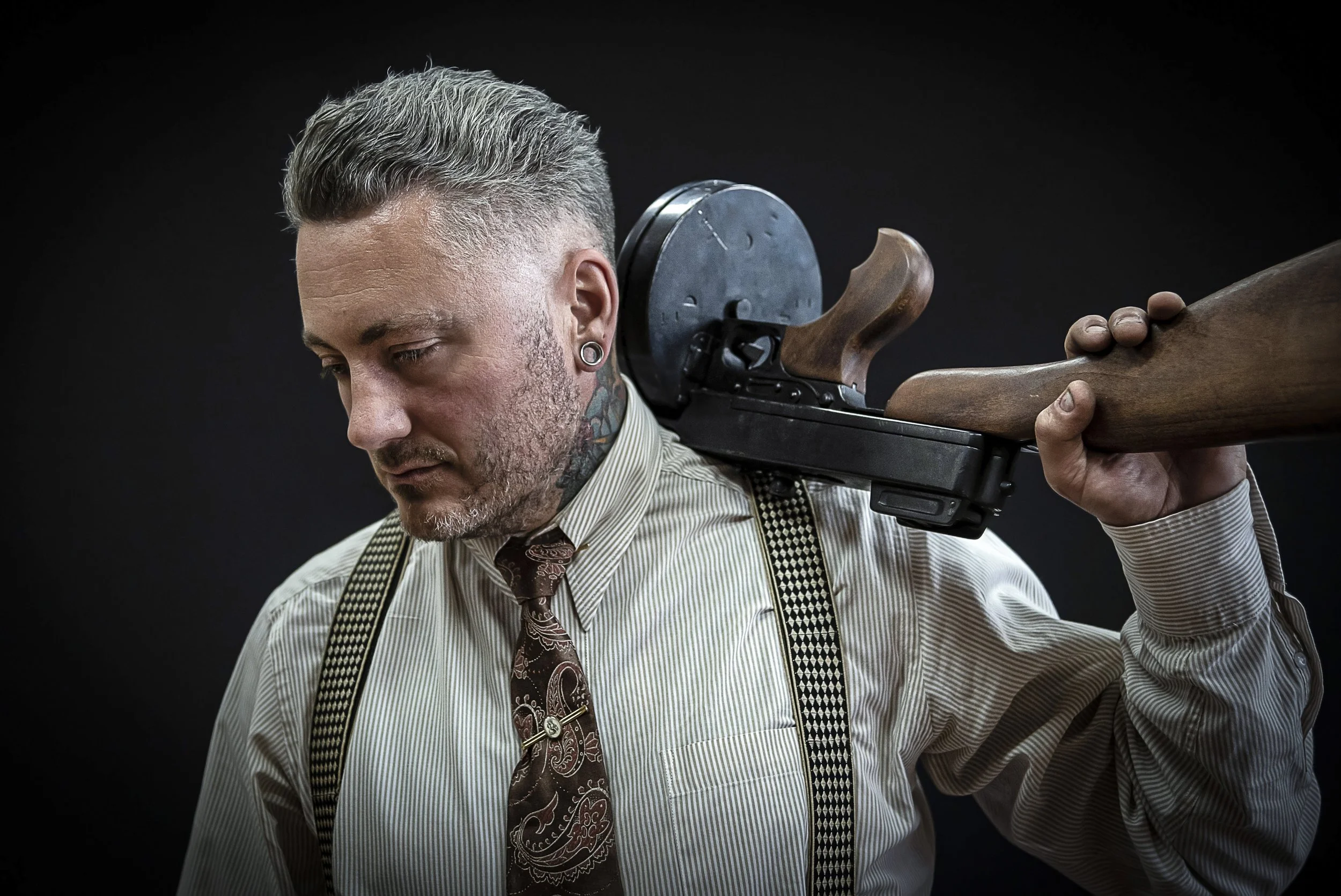 A man with short, gray hair and tattoos on his neck and ears, wearing a striped shirt, suspenders, and a paisley tie, holds a vintage rifle with a wooden stock over his shoulder against a dark background.