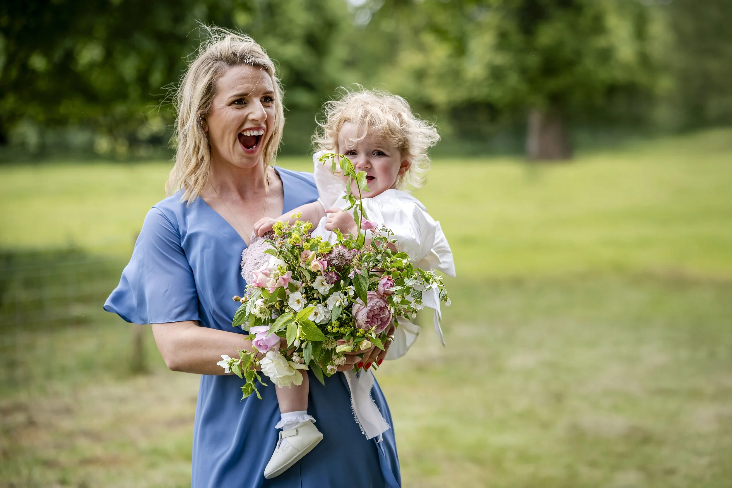 A woman in a blue dress holding a young child with blonde curly hair, outdoors in a green park, both smiling and surrounded by flowers.