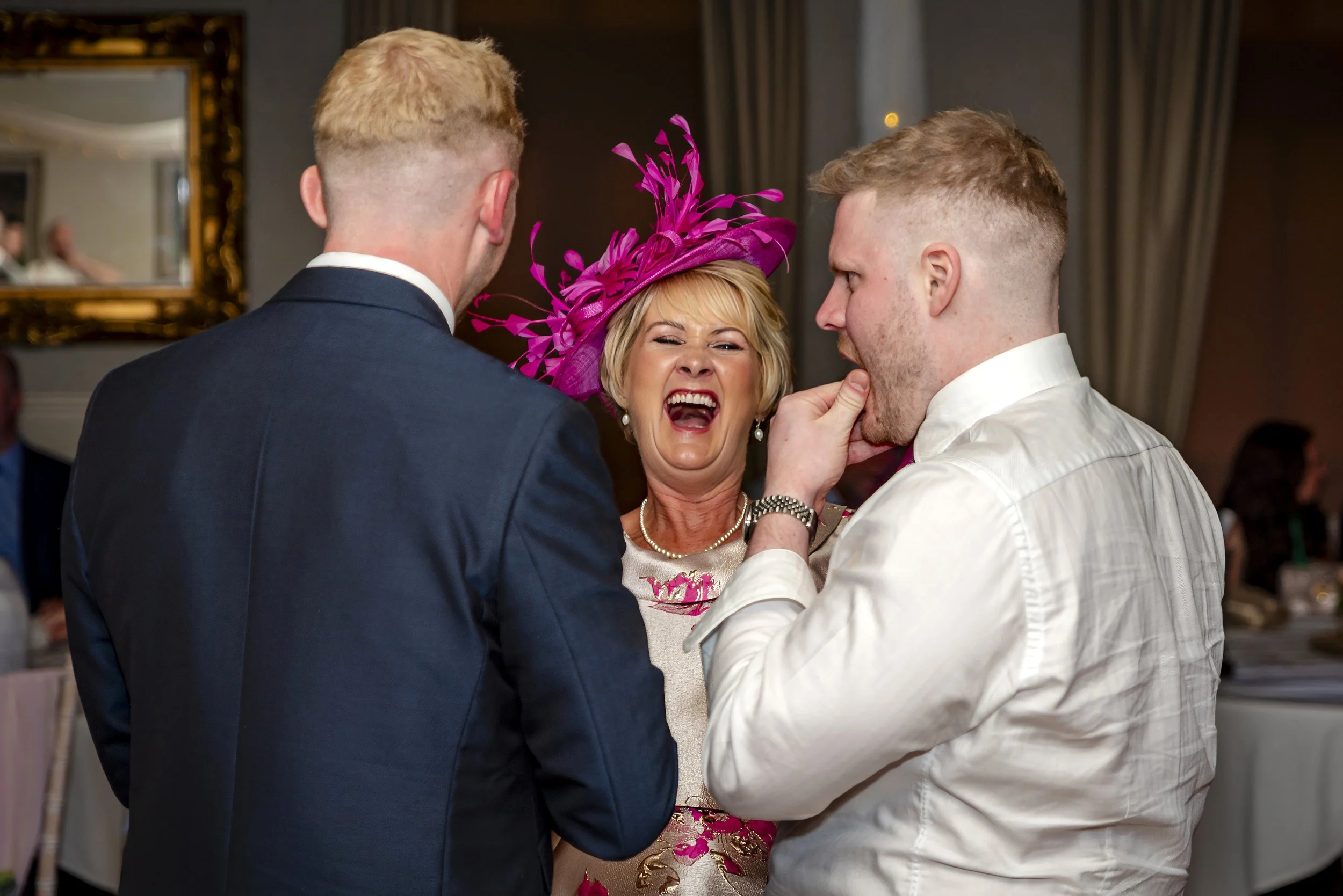 A woman wearing a bright pink hat laughing with her mouth open, as two men, one in a dark suit and the other in a white shirt, stand close to her and have a playful moment.