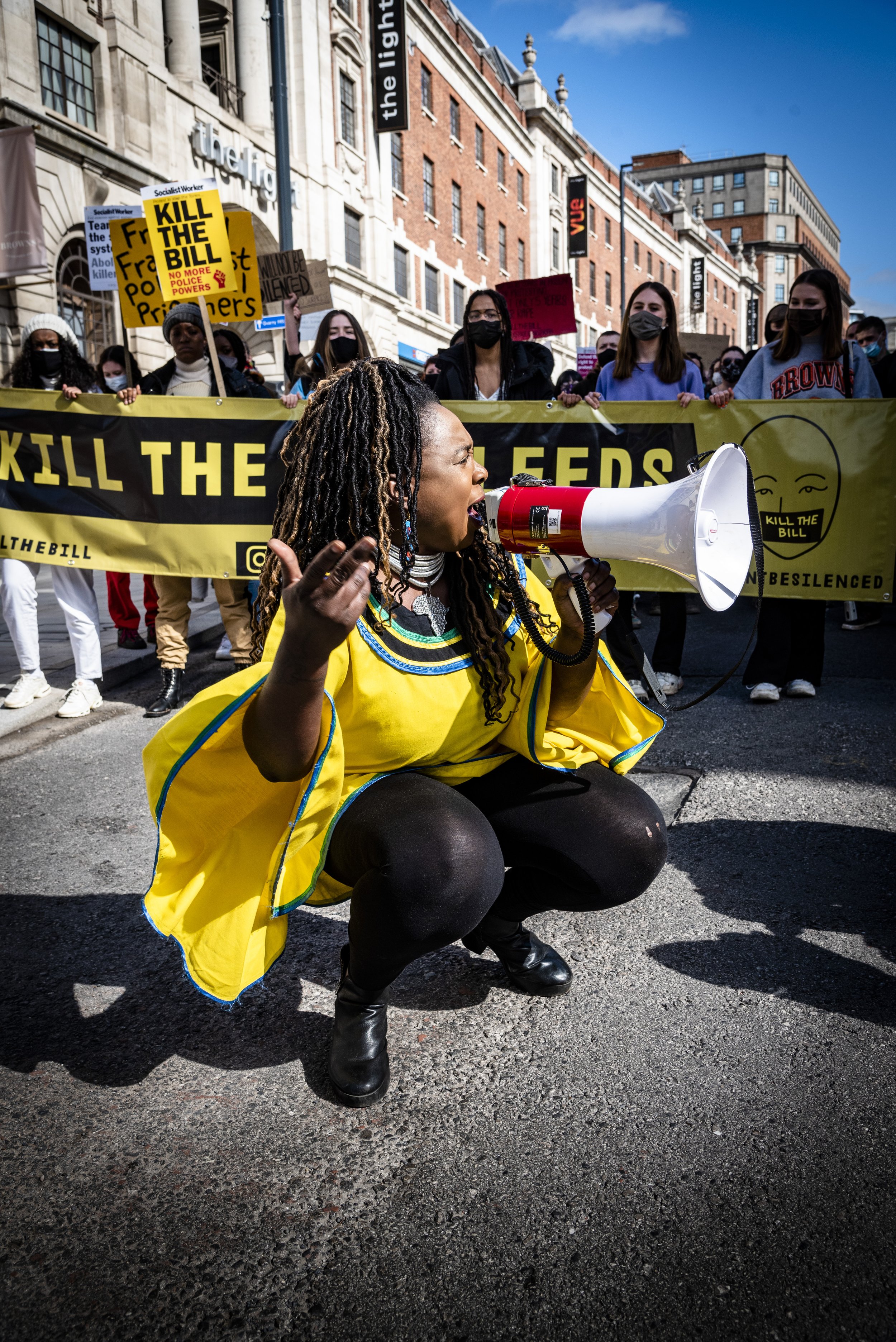 A woman kneeling on the street, holding a megaphone, participating in a protest. She is dressed in a bright yellow garment with blue accents and black leggings. Behind her, a large group of people, some holding signs that say "Kill the Bill" and othe