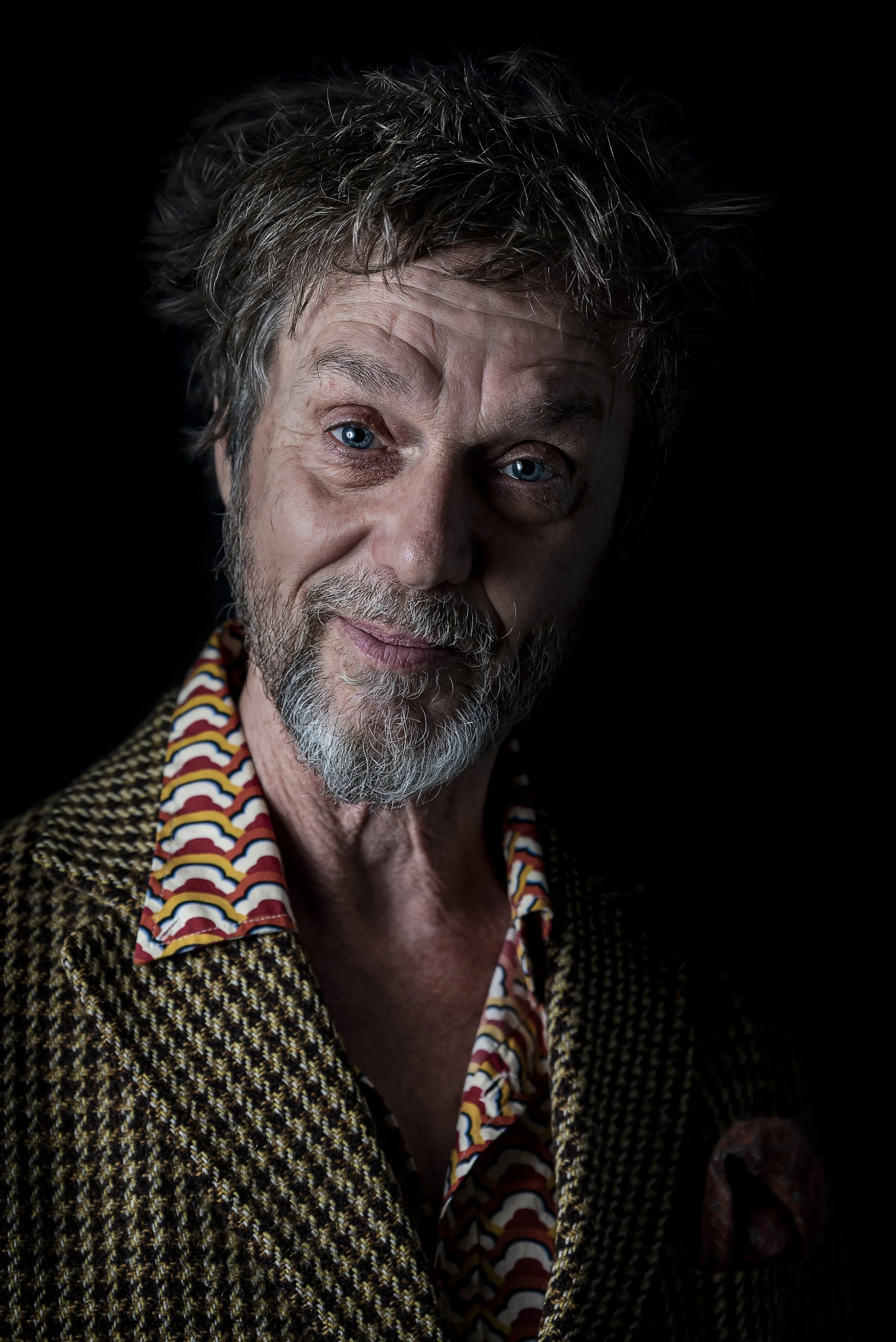 An elderly man with gray hair, blue eyes, and a beard, wearing a patterned shirt and a woven jacket, against a dark background.