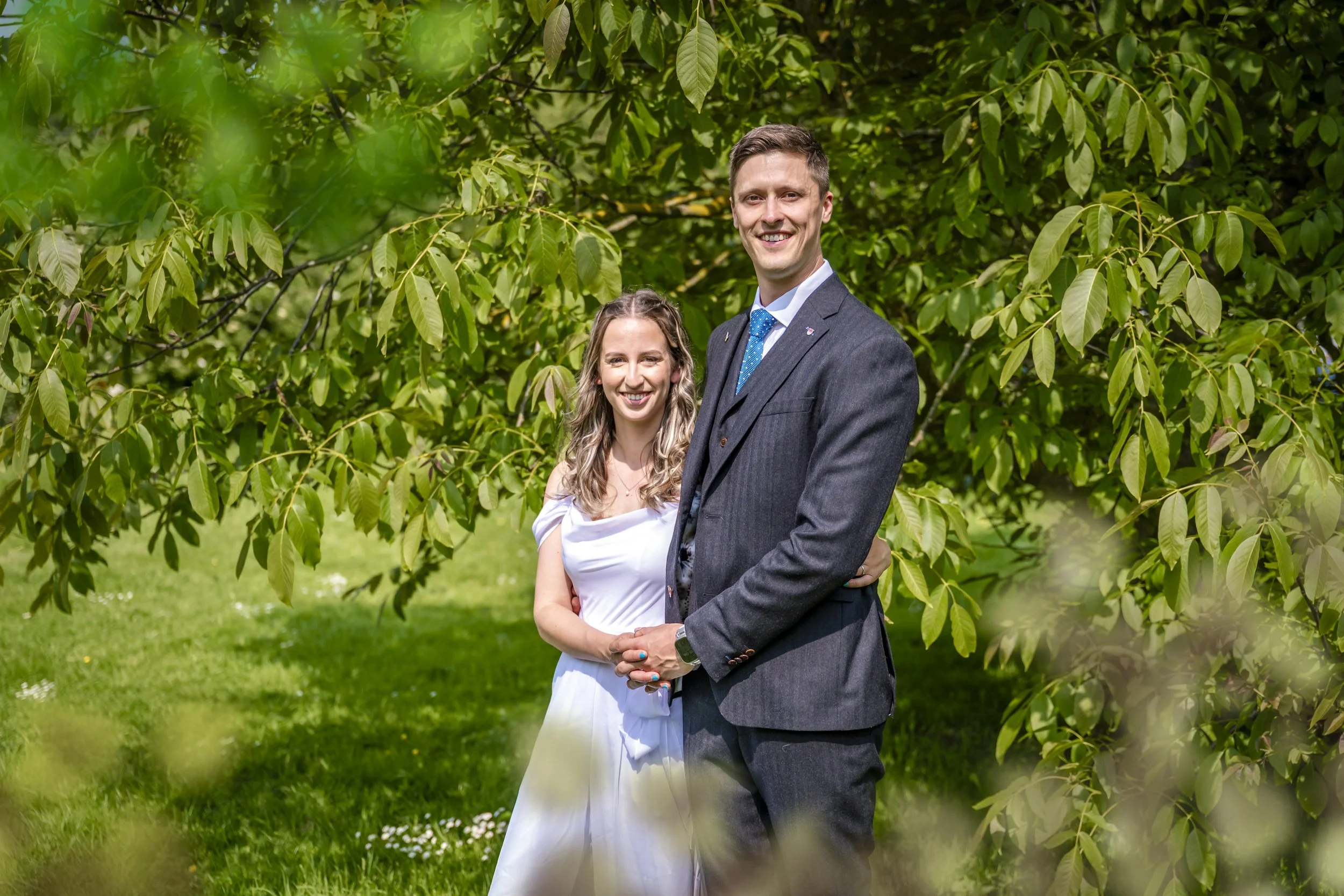 A smiling man and woman standing together outdoors among green trees and grass, dressed in formal attire.