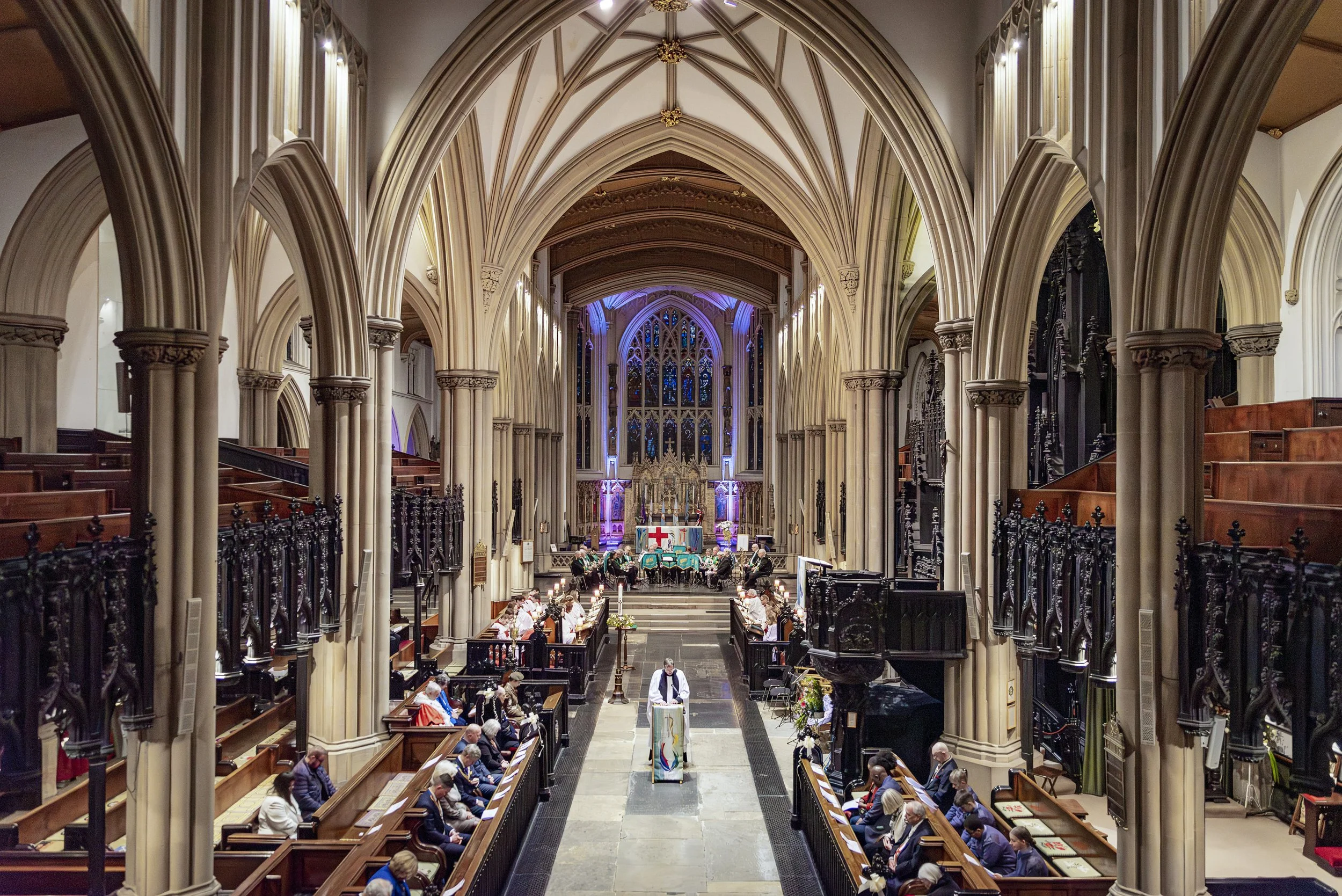 Interior of a grand Gothic-style church with high vaulted ceilings, stained glass windows, and ornate architecture. People are seated in wooden pews, and a choir or group of musicians is performing at the altar.