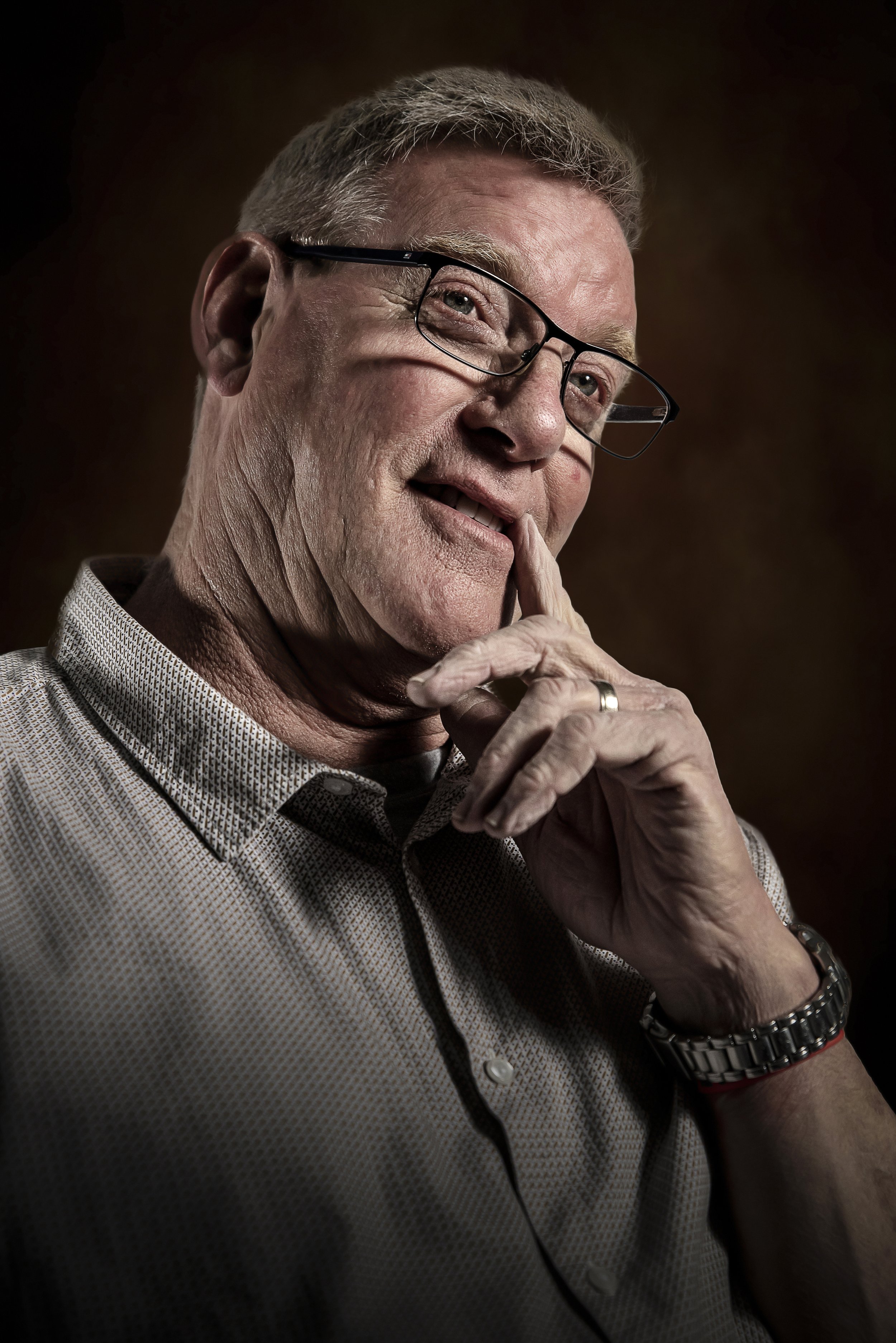 Close-up portrait of an elderly man with gray hair, wearing glasses and a patterned button-up shirt, touching his face thoughtfully.