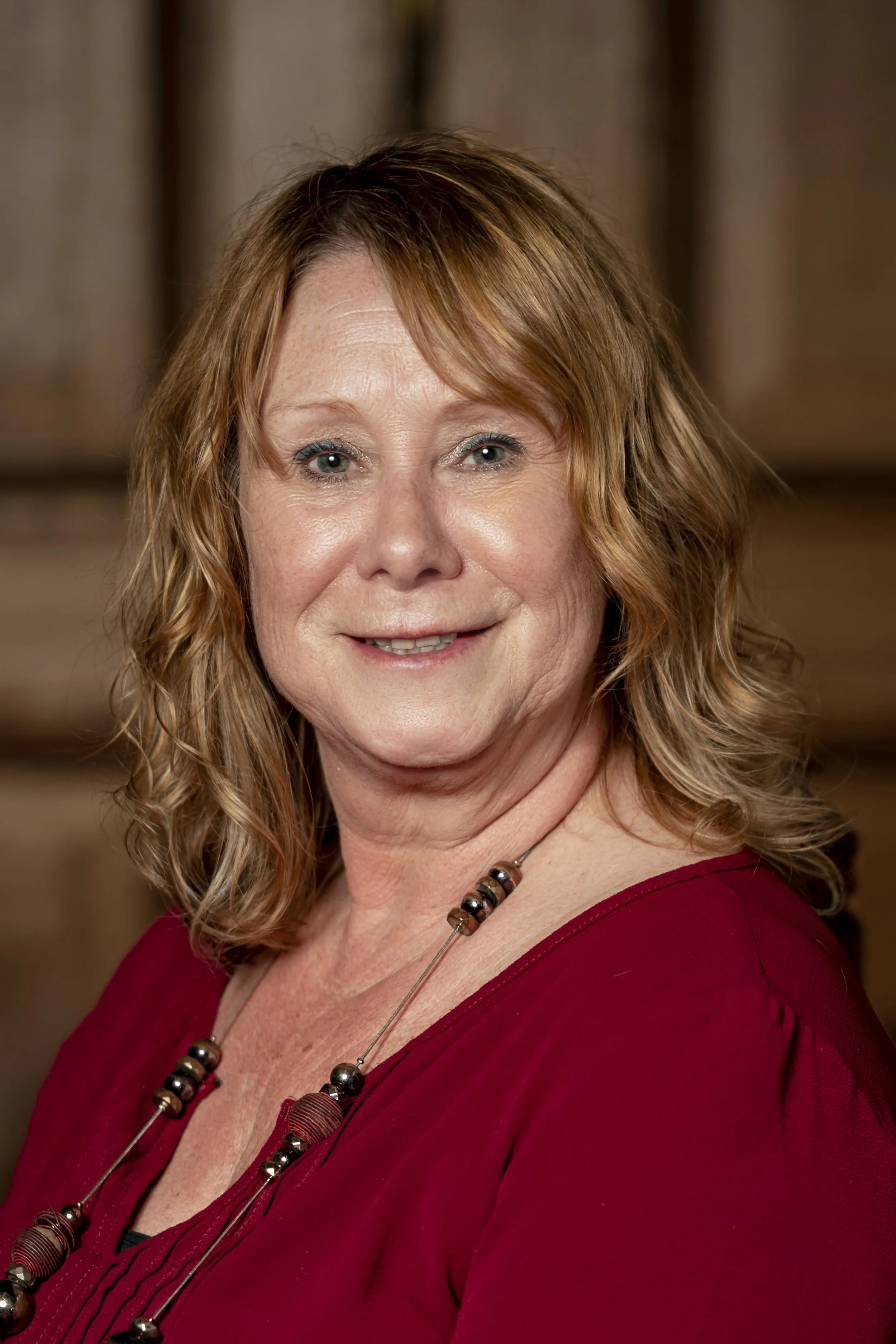 A smiling middle-aged woman with wavy red hair, wearing a red top and a beaded necklace, indoors with a wooden background.