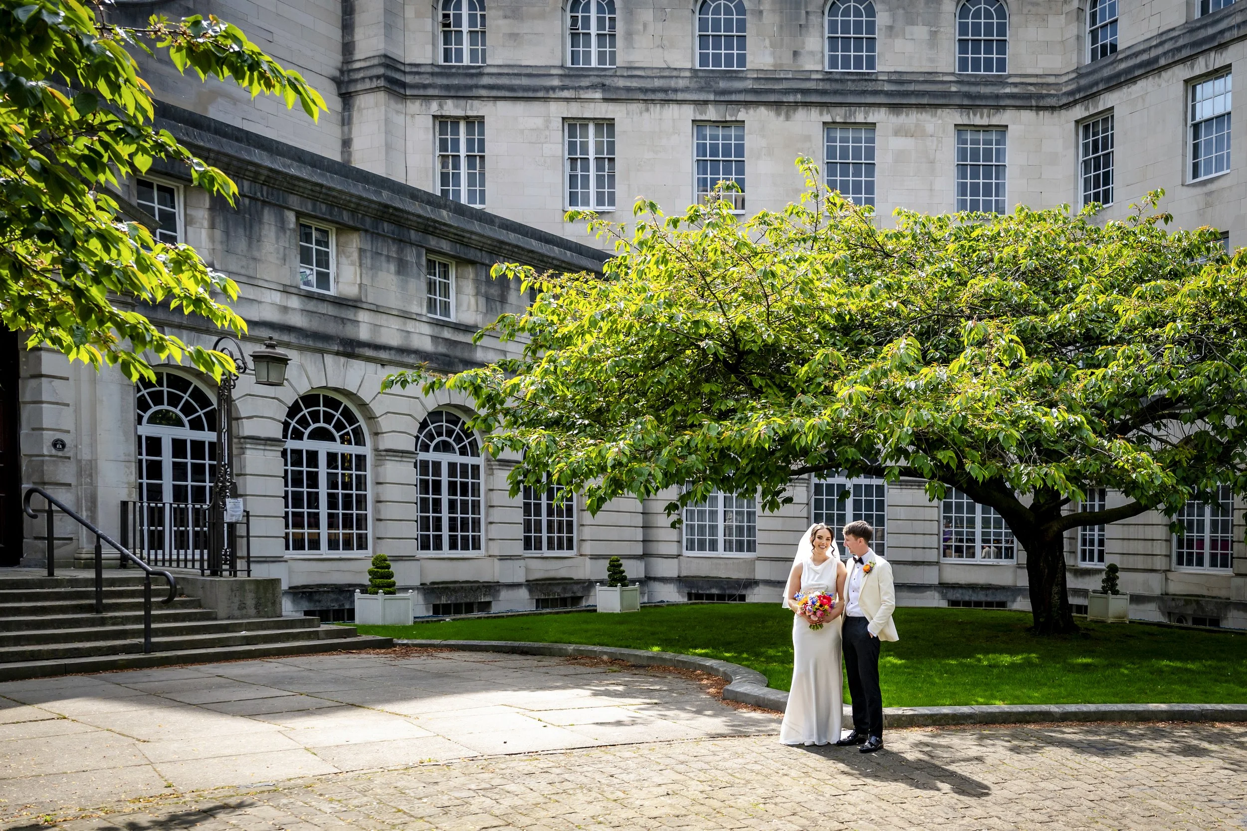 A bride and groom standing outdoors in front of a large stone building with multiple windows and a rounded corner, trees, and stairs. The bride is holding a bouquet of flowers, and they are smiling at each other.