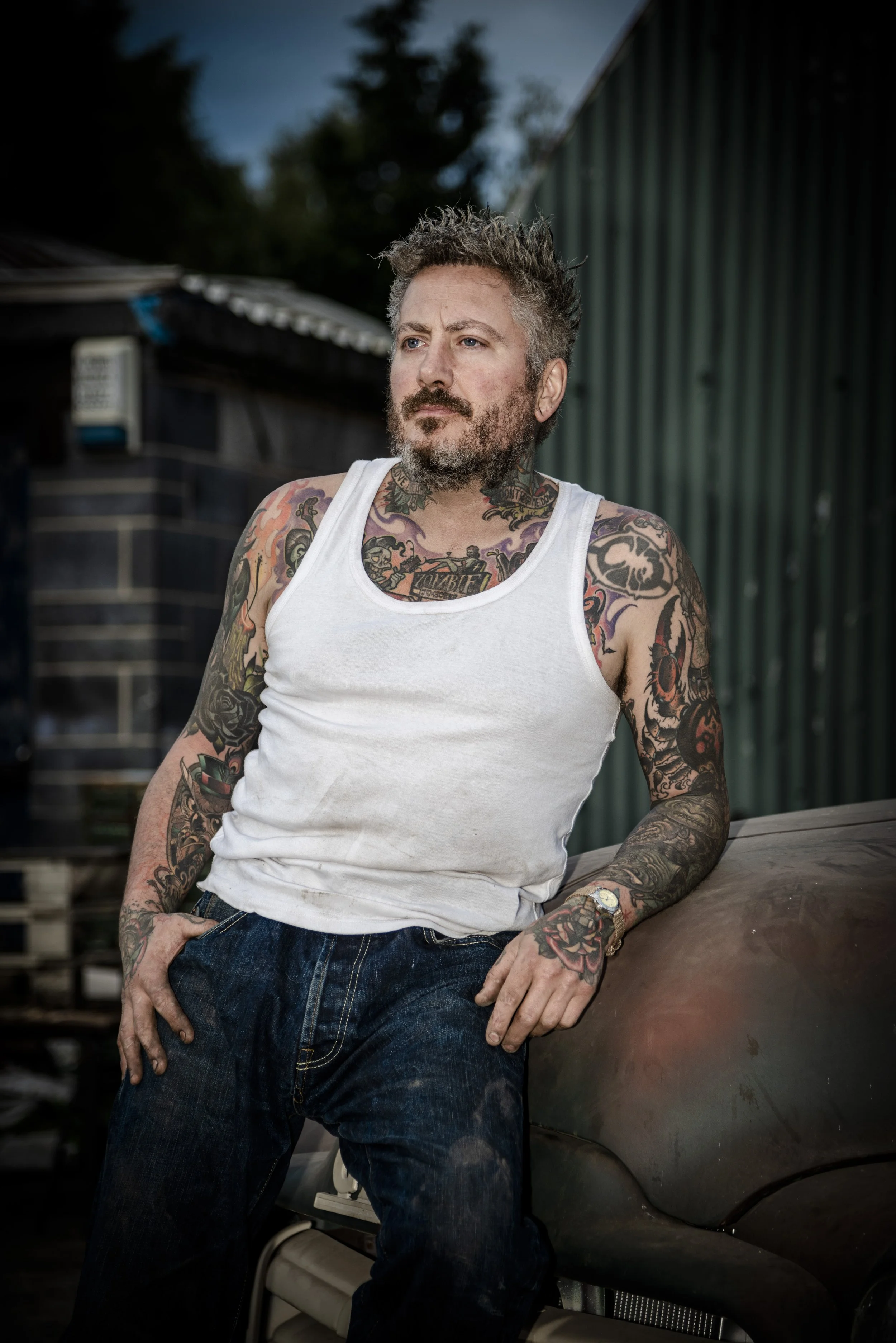 Tattooed man in white tank top and jeans standing outdoors near a vintage car.
