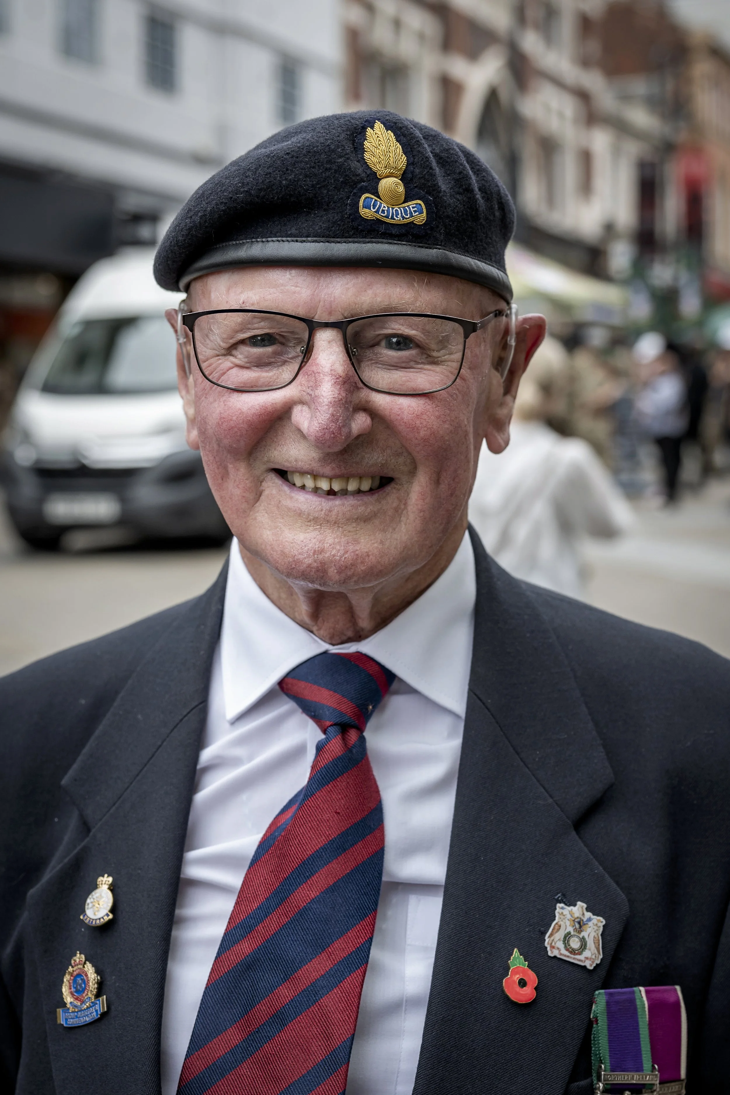 An elderly man wearing a black beret with a golden badge, glasses, a white shirt, a suit jacket with military medals and ribbons, and a striped tie. He is smiling and standing outdoors on a busy street.