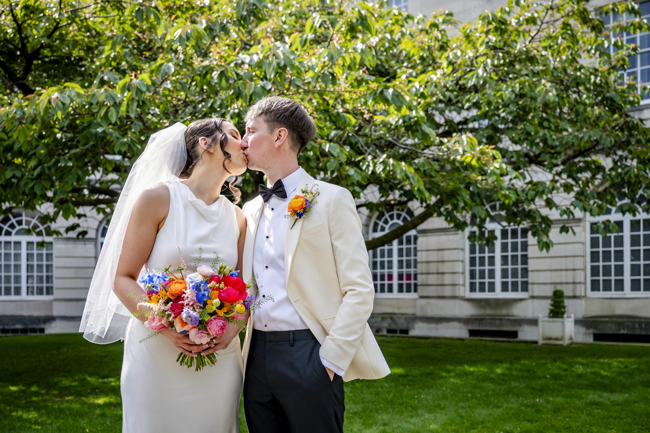 A newlywed couple sharing a kiss outdoors, with the bride holding a colorful bouquet of flowers and wearing a white wedding dress with a veil, and the groom dressed in a white tuxedo with a black bow tie, standing in front of green trees and a histor