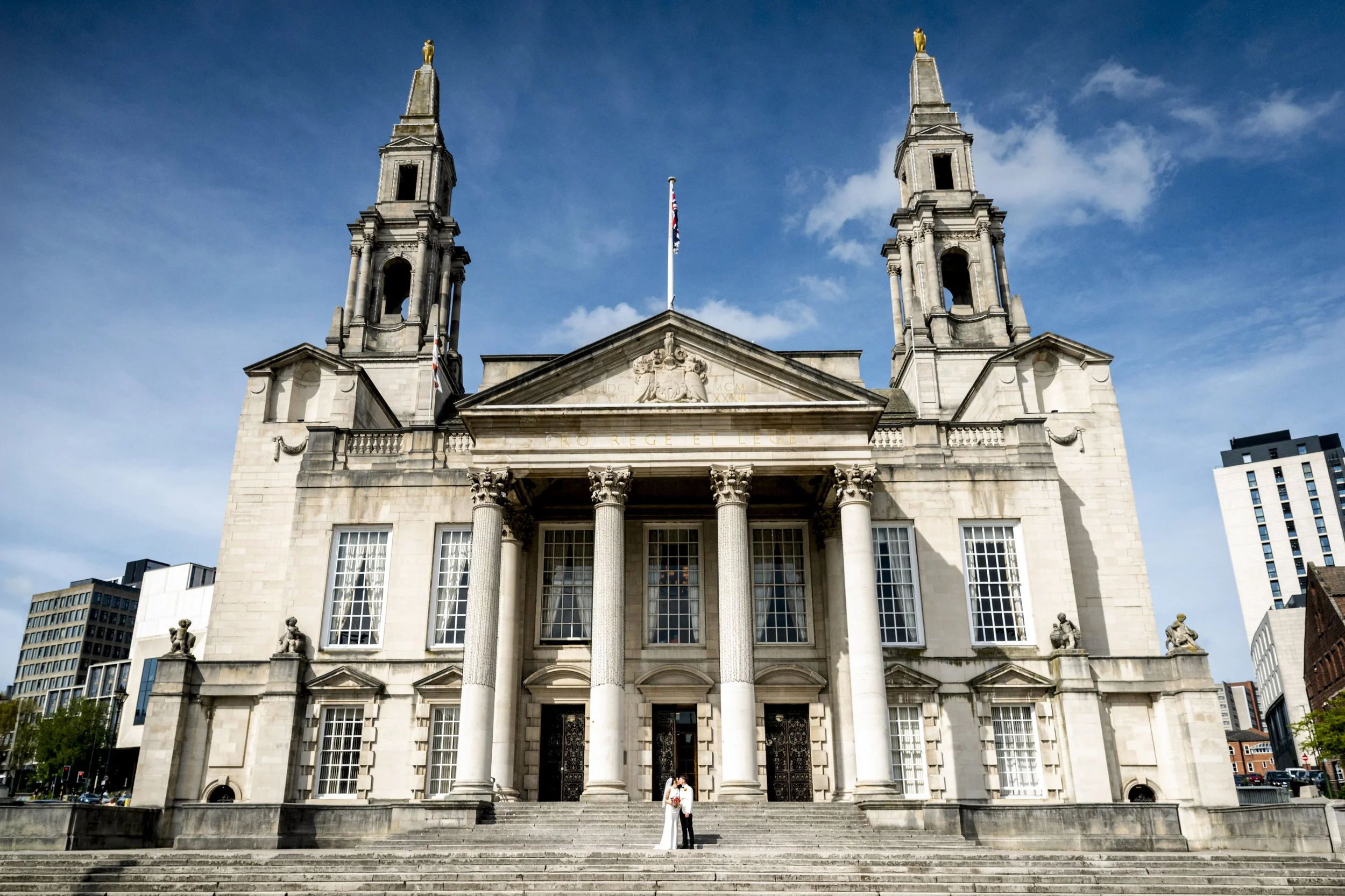 A grand, historic building with classical architecture, featuring two tall towers with spires, a triangular pediment with a crest, and large windows. A British flag is flying in front of the building, and two people are standing on the steps below.