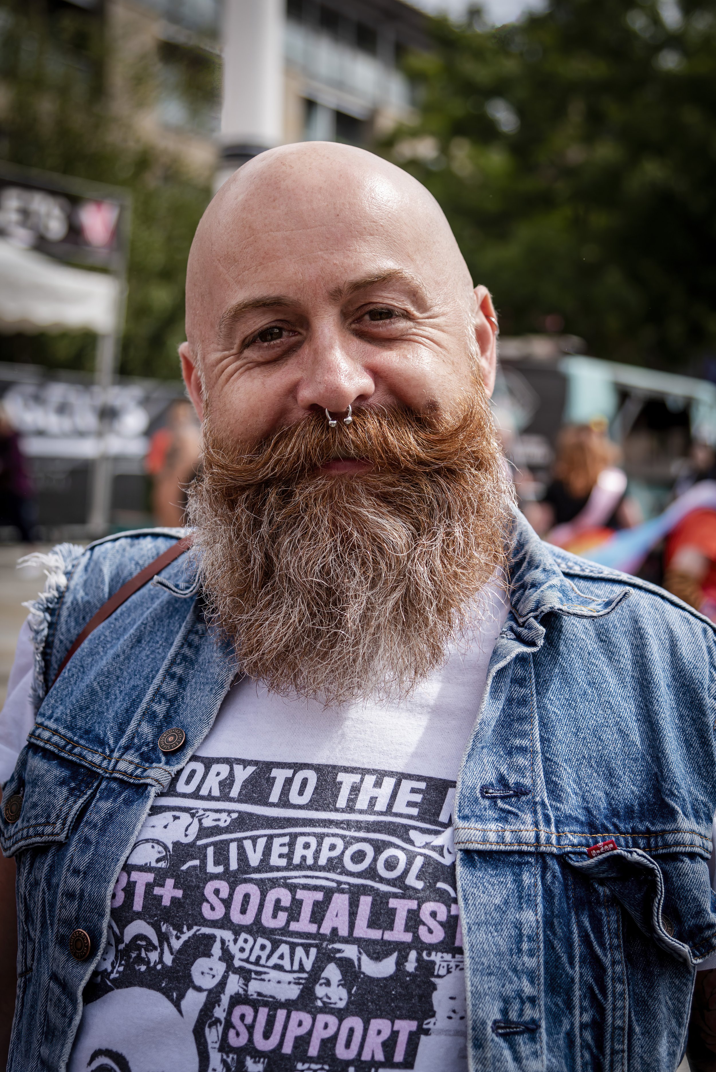 A man with a bald head and a large, full beard smiling outdoors at a festival or gathering, wearing a denim vest and a graphic T-shirt.