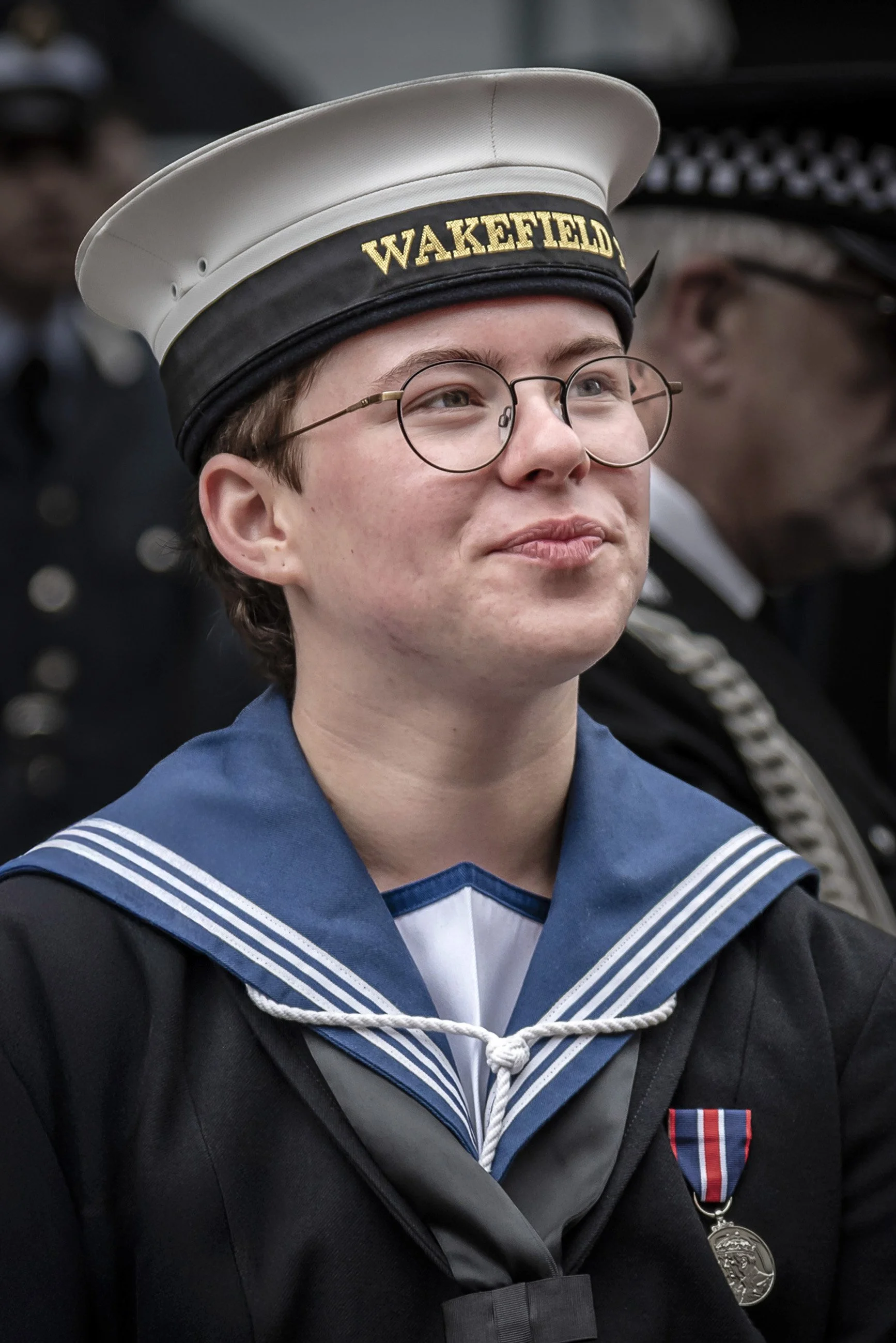A young man wearing a navy sailor uniform with a white hat that has 'Wakefield' embroidered on it, glasses, and a medal on his chest, standing among other uniformed individuals in a formal setting.
