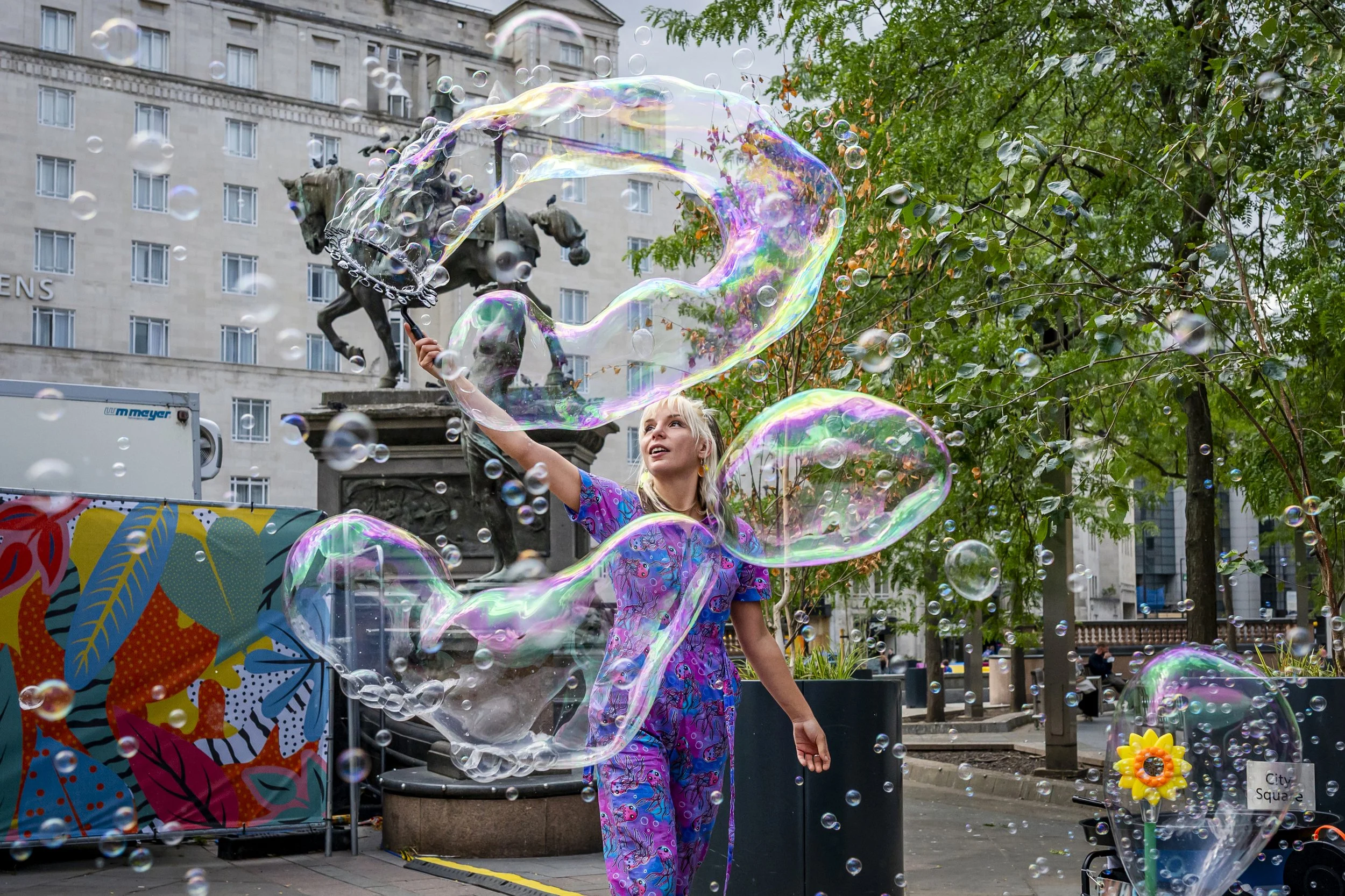 A woman in colorful pajamas is creating large soap bubbles in an urban park with trees and buildings in the background. There is a statue of a mounted figure nearby and a display with a colorful leaf pattern.