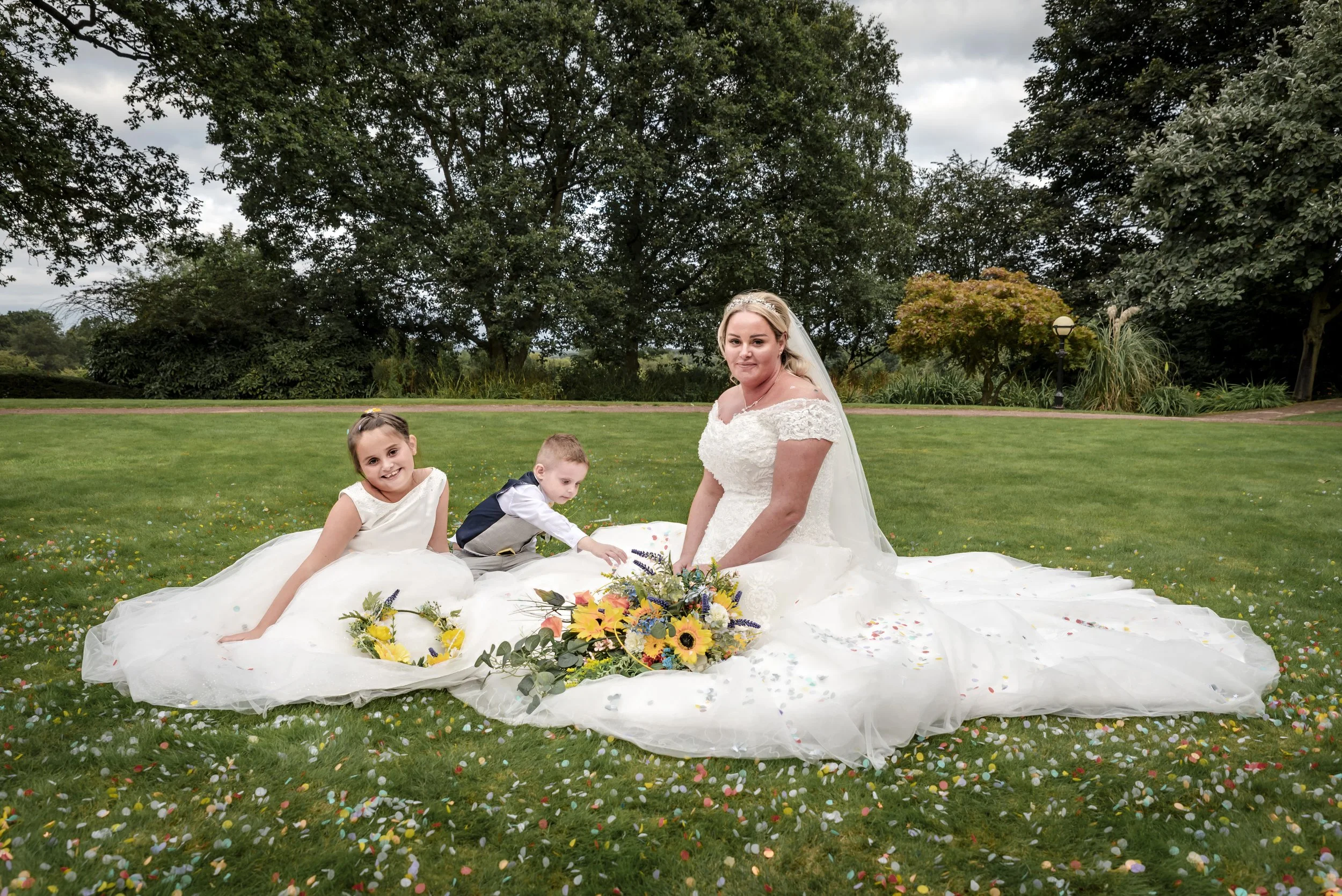 Bride in a white wedding gown sitting on the grass with two children, surrounded by confetti and flowers, in an outdoor park with trees and cloudy sky in the background.