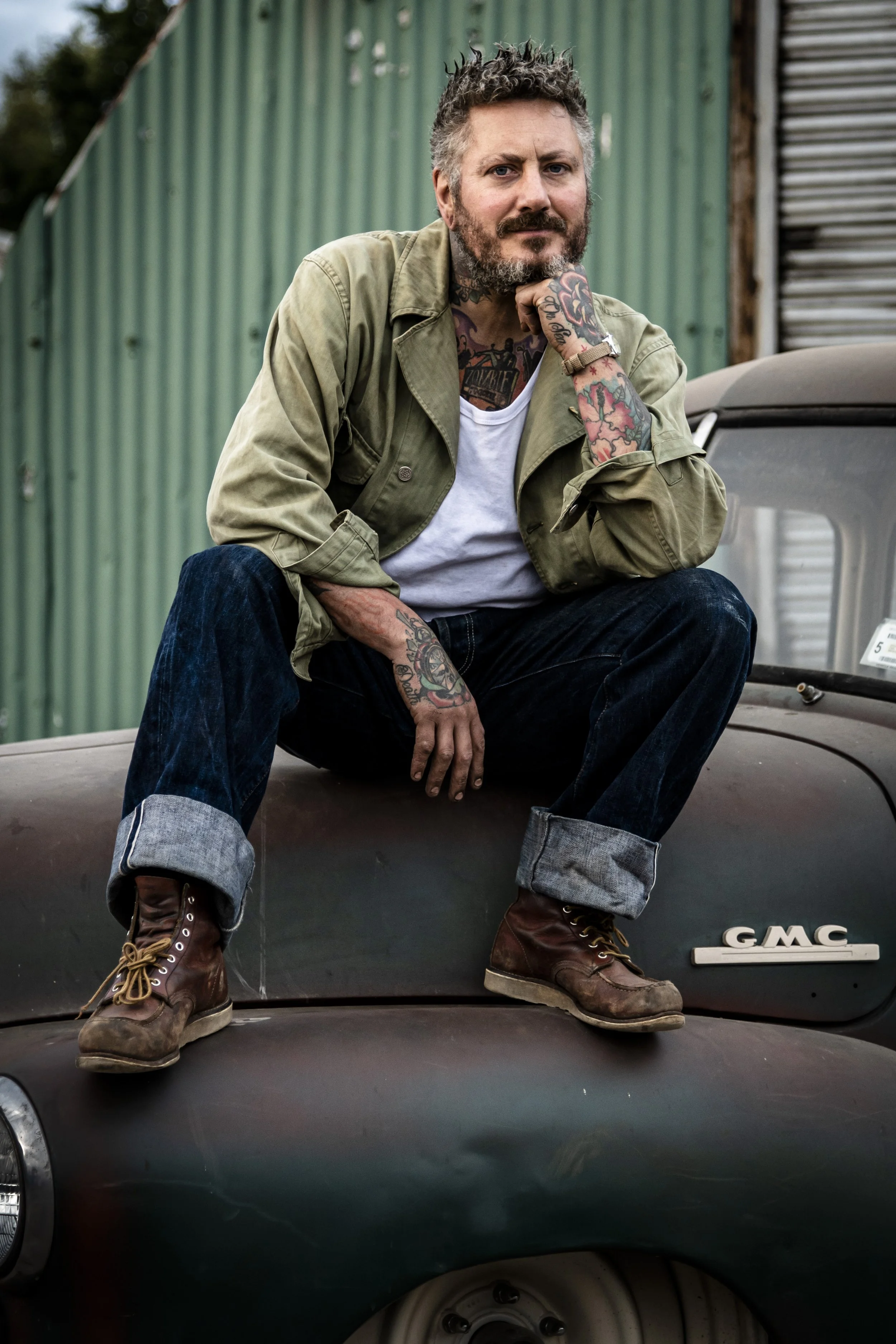 A bearded man with tattoos, wearing a green jacket, white t-shirt, dark jeans, and brown boots, sitting on the hood of an old GMC truck with a green and gray building in the background.