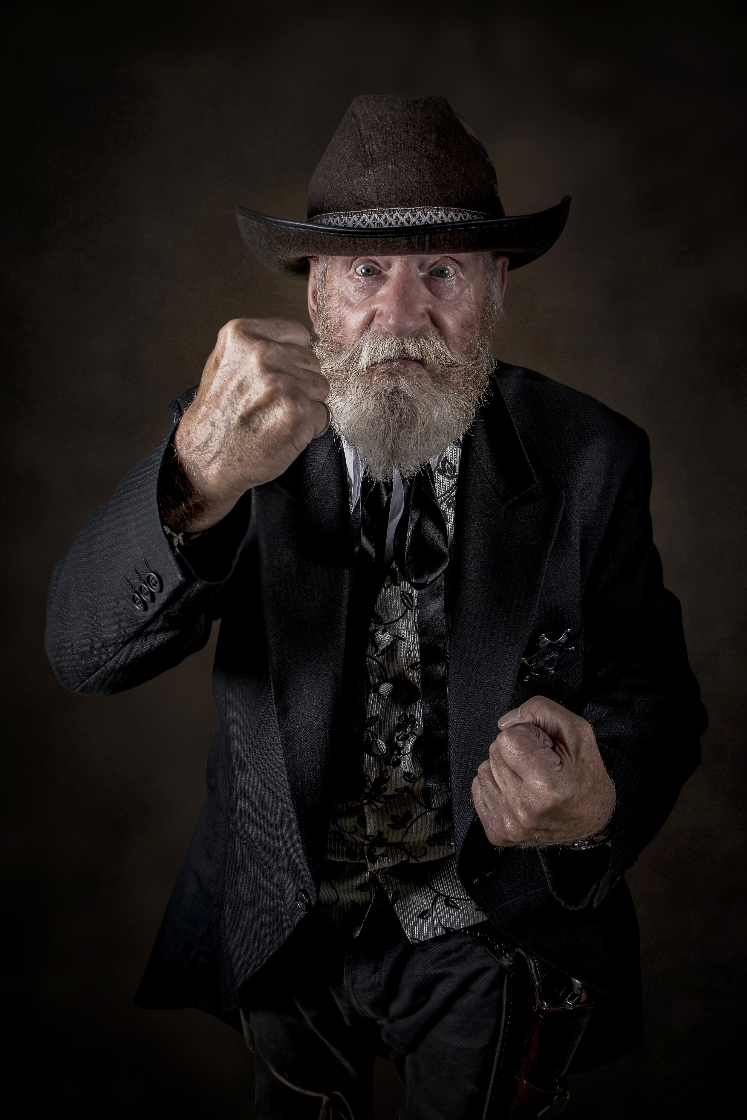 An elderly man with a gray beard and mustache wearing a dark cowboy hat, a dark suit, and a patterned shirt, showing a fierce expression with clenched fists.