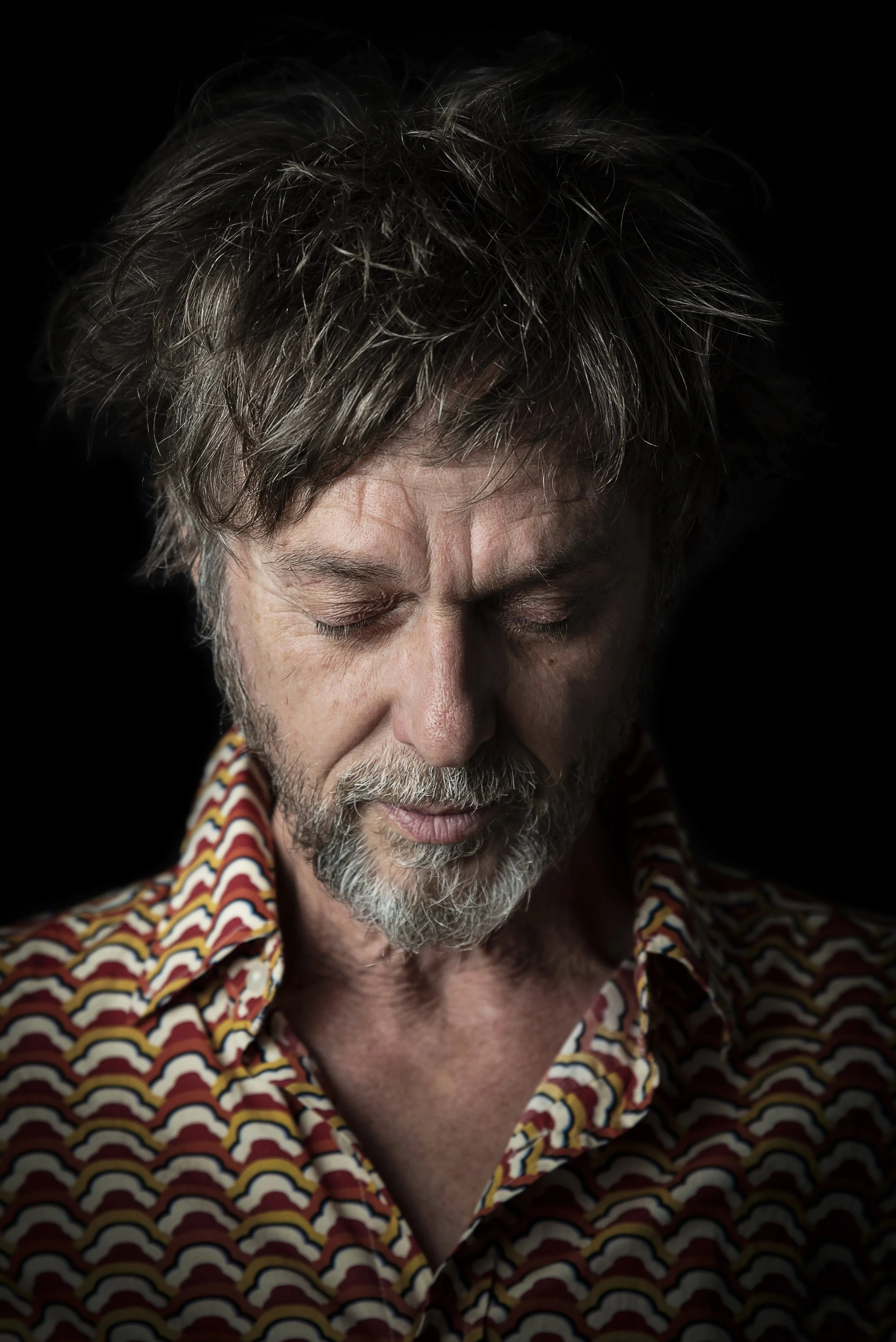 A man with disheveled gray hair and beard, eyes closed, wearing a patterned shirt, against a black background.