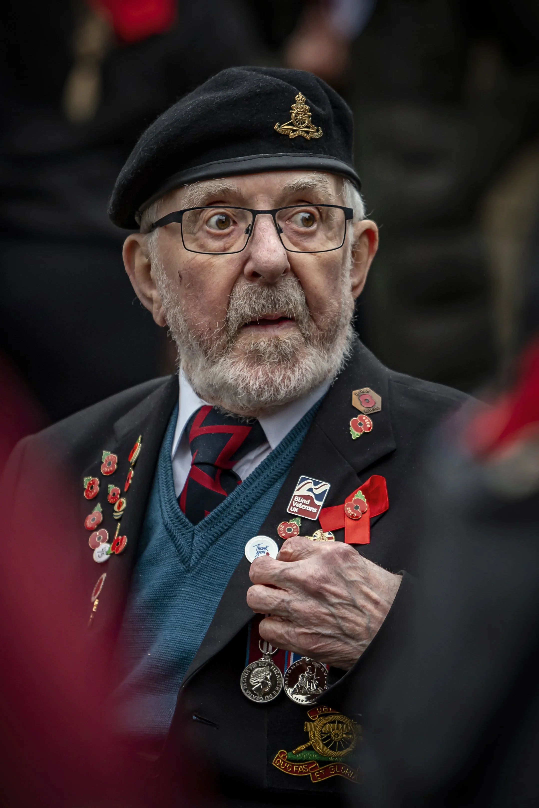 An elderly man with glasses, a beard, and a black beret with a badge is wearing a suit jacket decorated with poppy pins, military medals, and a badge that says 'Blind Veterans UK.' He is holding a lapel pin with his right hand and has a surprised or 