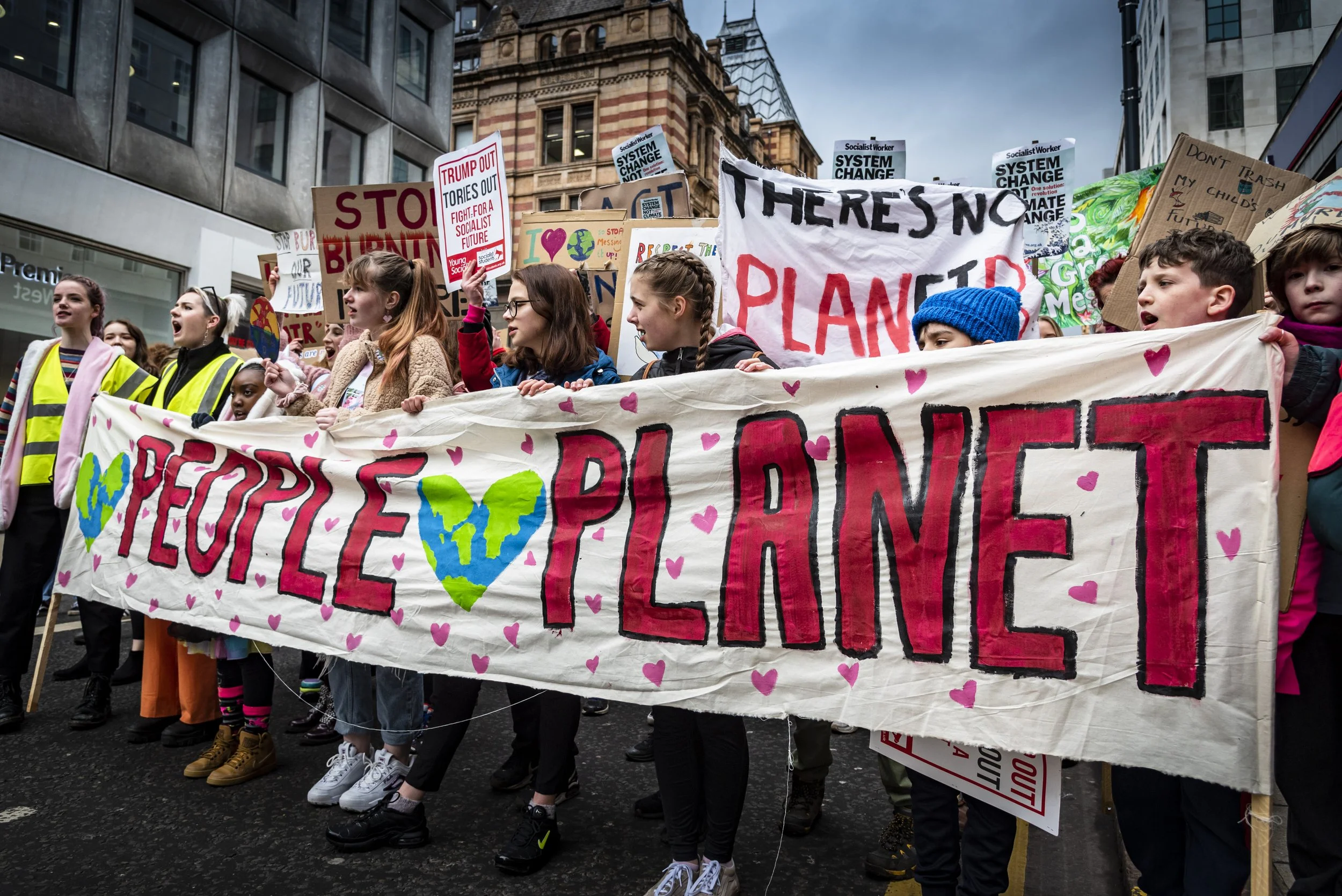 A large group of young protesters marching in the city streets, holding various signs and banners advocating for climate action and social justice. Some signs read 'STOP BURNING OUR FUTURE,' 'THERE'S NO PLANET B,' and 'RESPECT THE EARTH.'