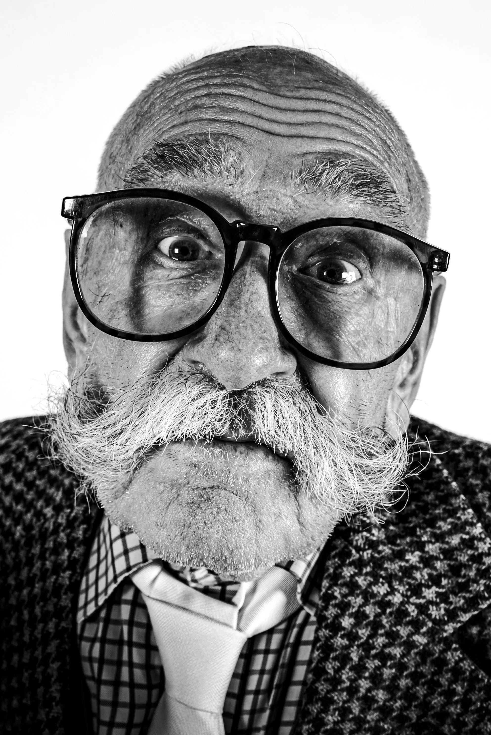 Close-up black and white portrait of an elderly man with glasses, a mustache, and a beard, wearing a checkered shirt, tie, and textured blazer, with a surprised or curious expression.