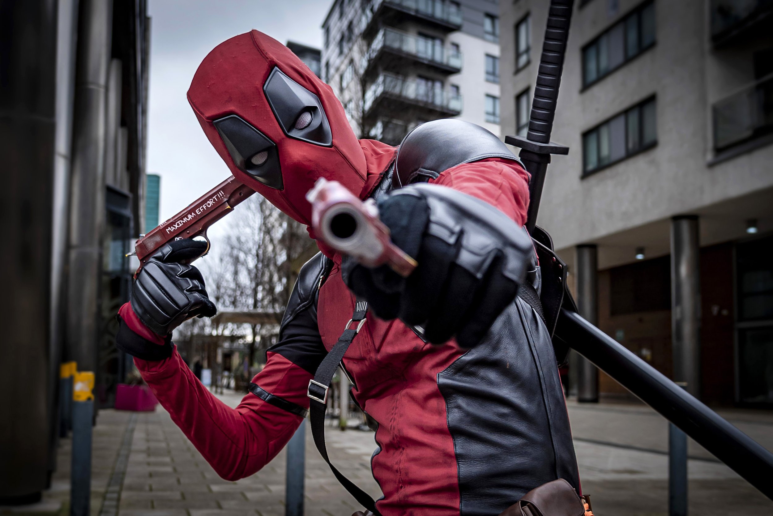 A person in a Deadpool mask and red jacket holding a katana sword outdoors in an urban setting with buildings and trees in the background.