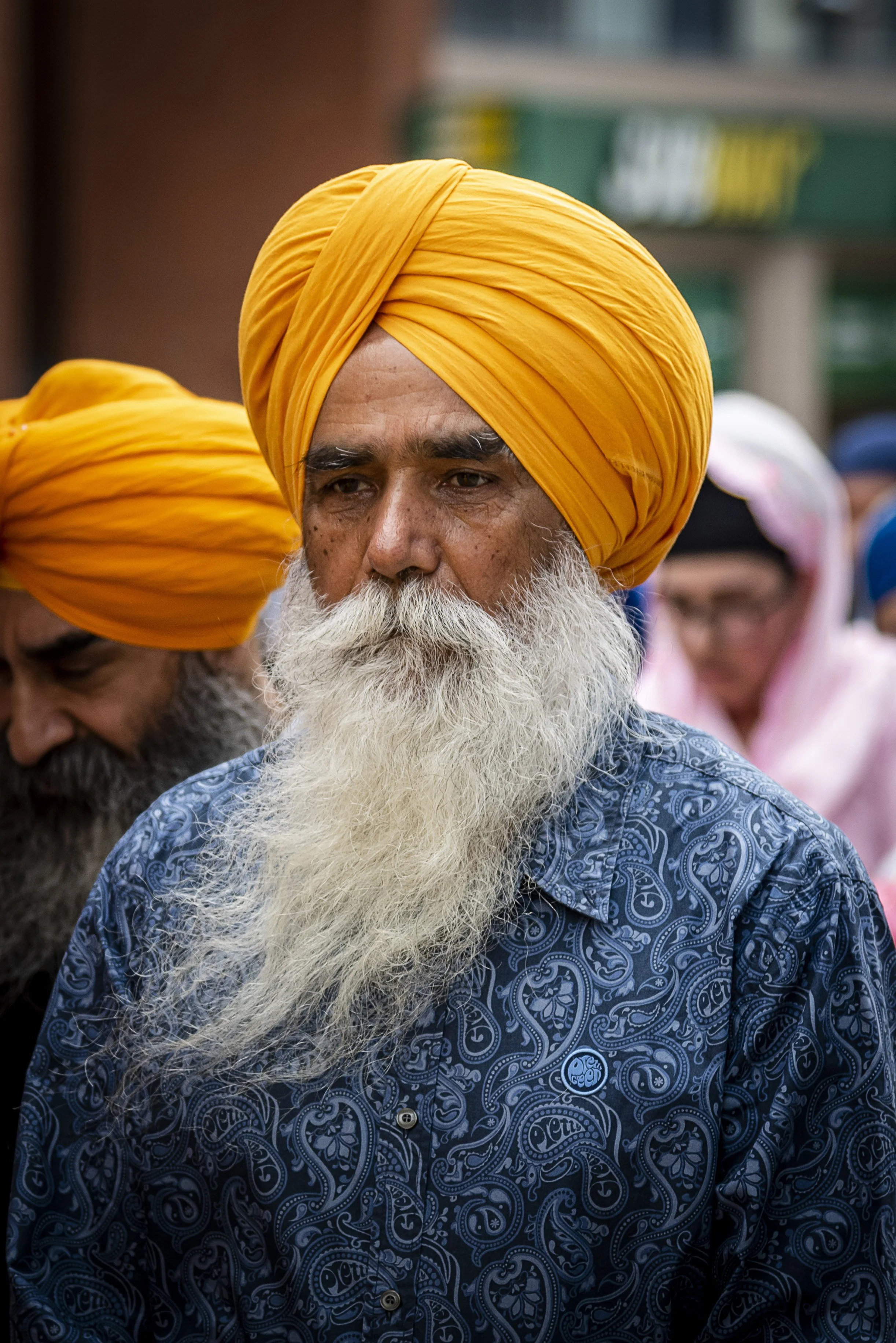 A man with a long white beard wearing a bright orange turban and a dark blue shirt with paisley pattern, standing among a group of people, some also wearing turbans and head coverings.