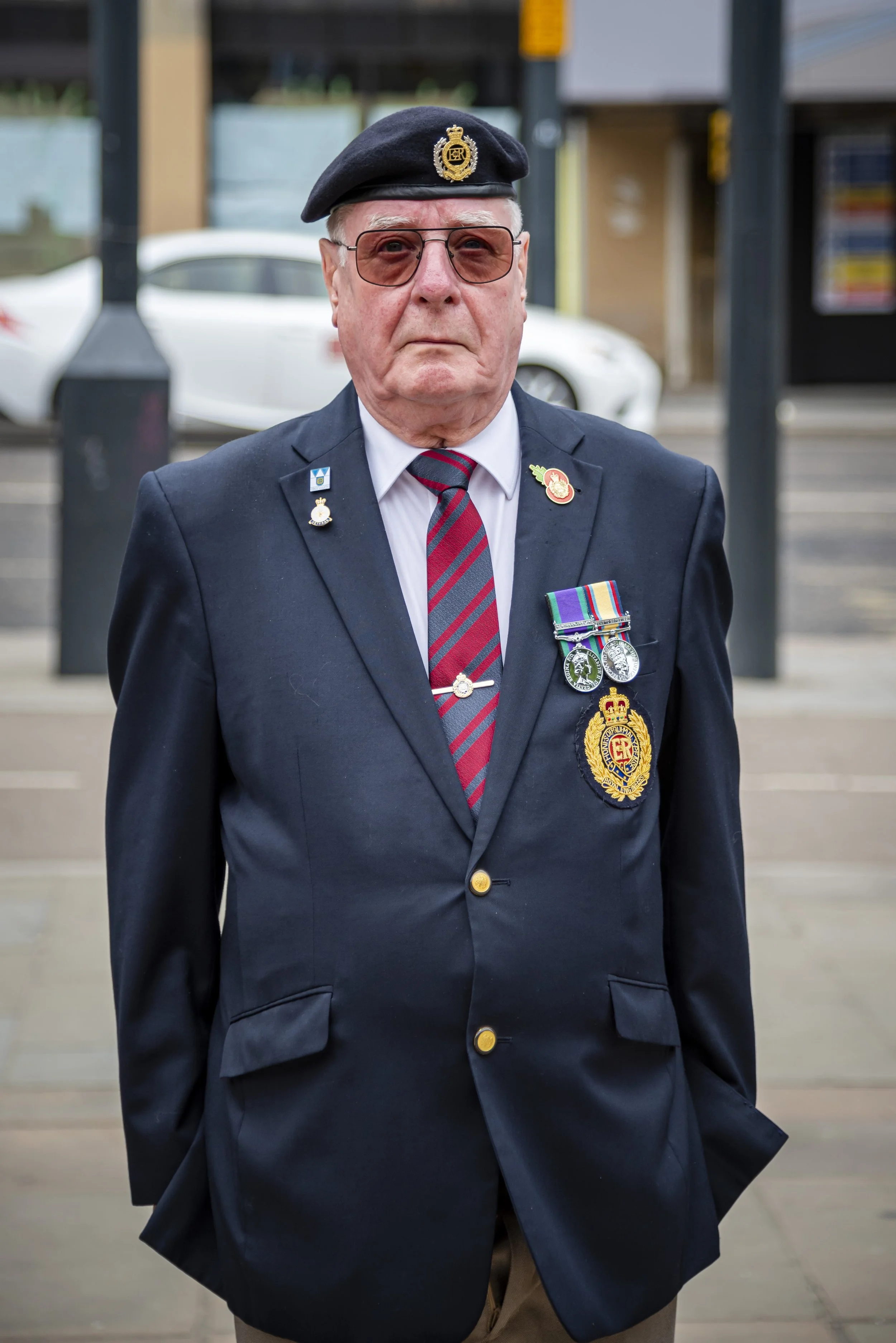 An elderly man wearing a dark blazer with military medals, a tie, a beret with insignia, sunglasses, and badges, standing outdoors near parked cars.