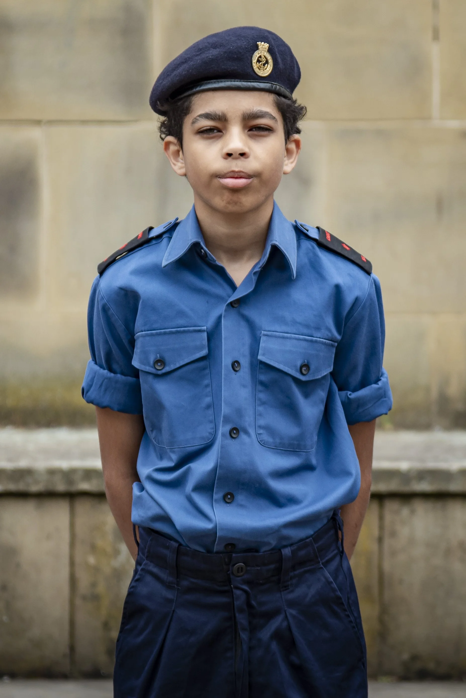 A young boy dressed in a blue police uniform with a hat featuring a badge, standing outdoors against a stone wall.