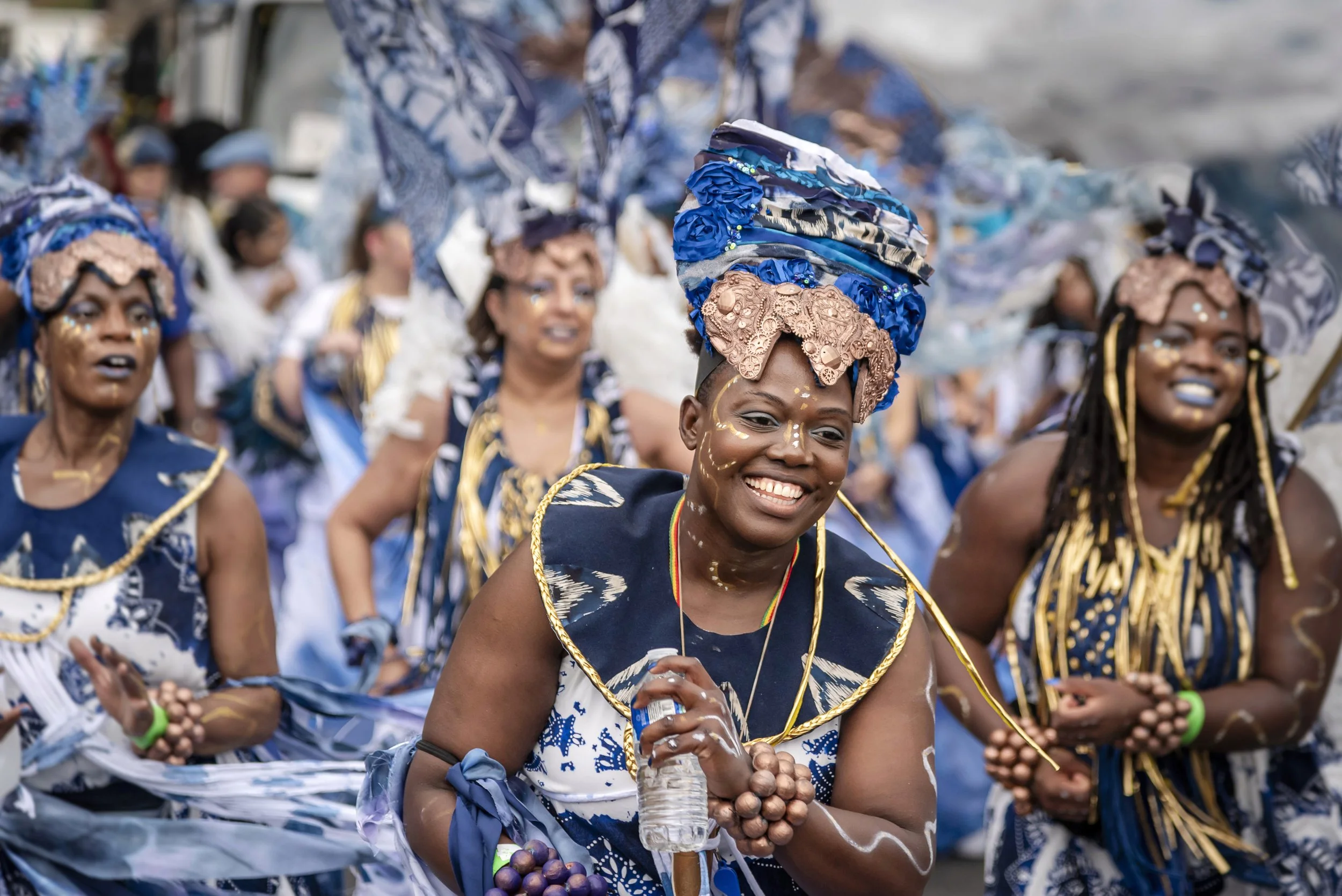 Women dressed in traditional clothing participating in a parade or festival, wearing elaborate headpieces and face paint, with a joyful expression.