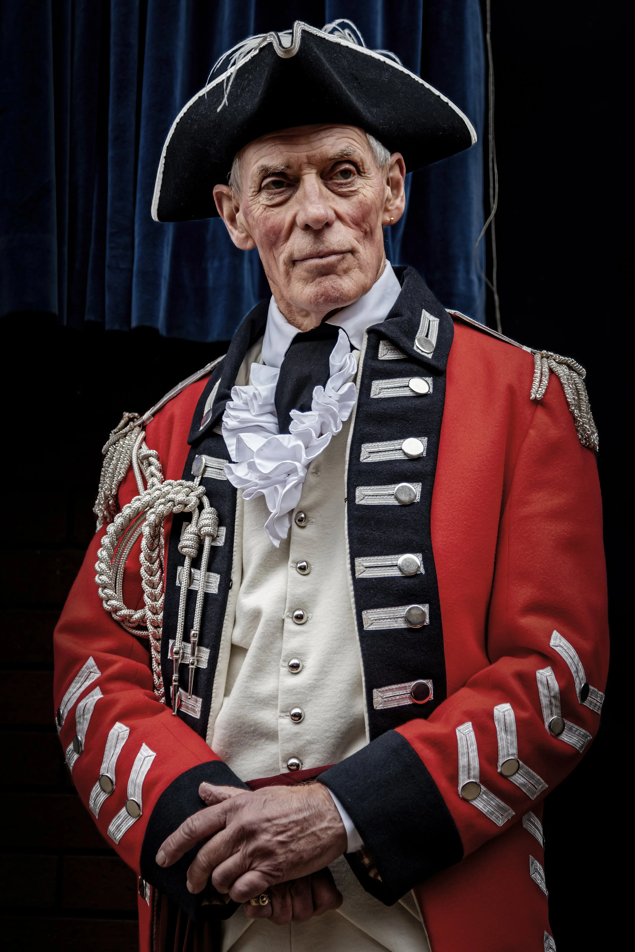 A man dressed in a historical military uniform with a red coat, white shirt, and black hat, standing in front of a dark background with blue curtains.