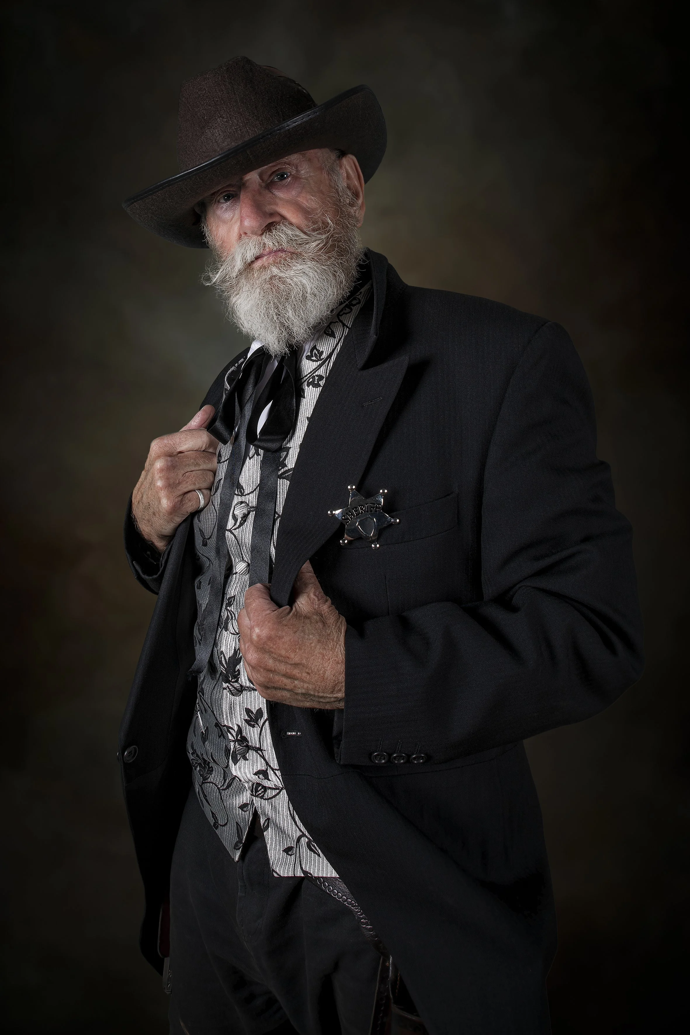 A man dressed as a cowboy sheriff wears a dark hat, a black suit with a sheriff badge, a patterned shirt, and a neckerchief, posing against a dark background.