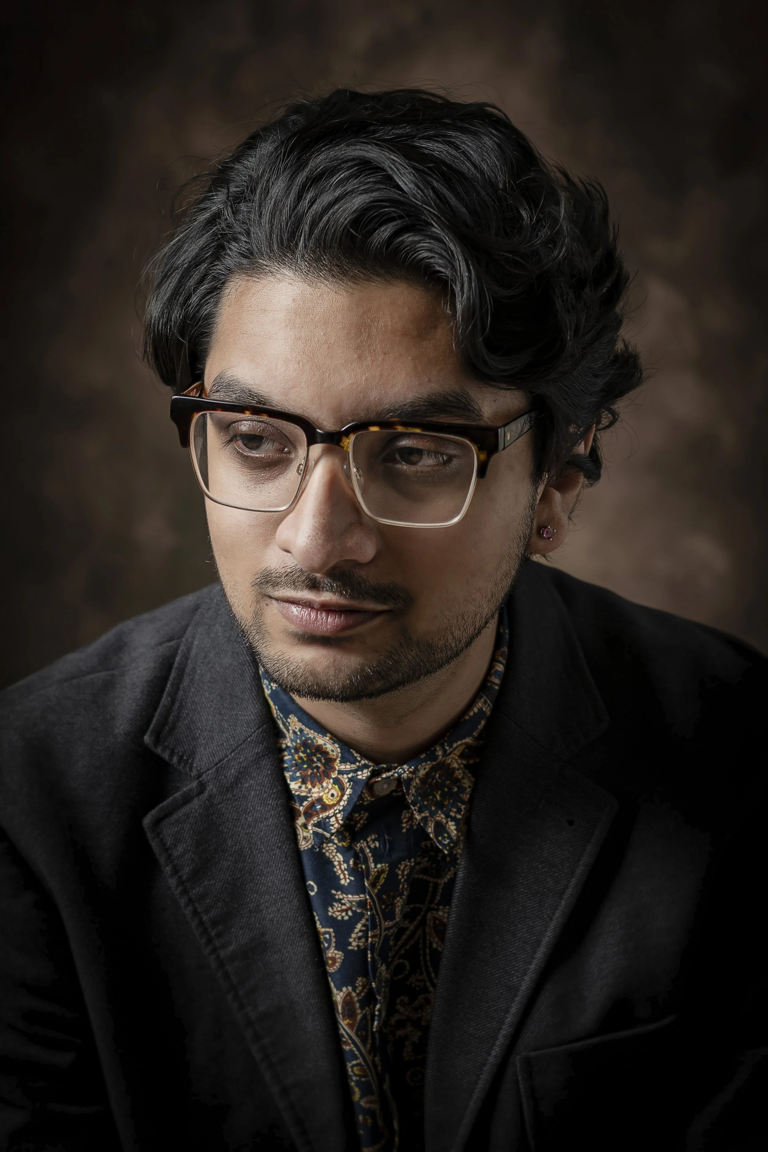 A young man with black wavy hair, glasses, and a light beard wearing a dark blazer over a patterned shirt, looking to the side against a brown blurred background.