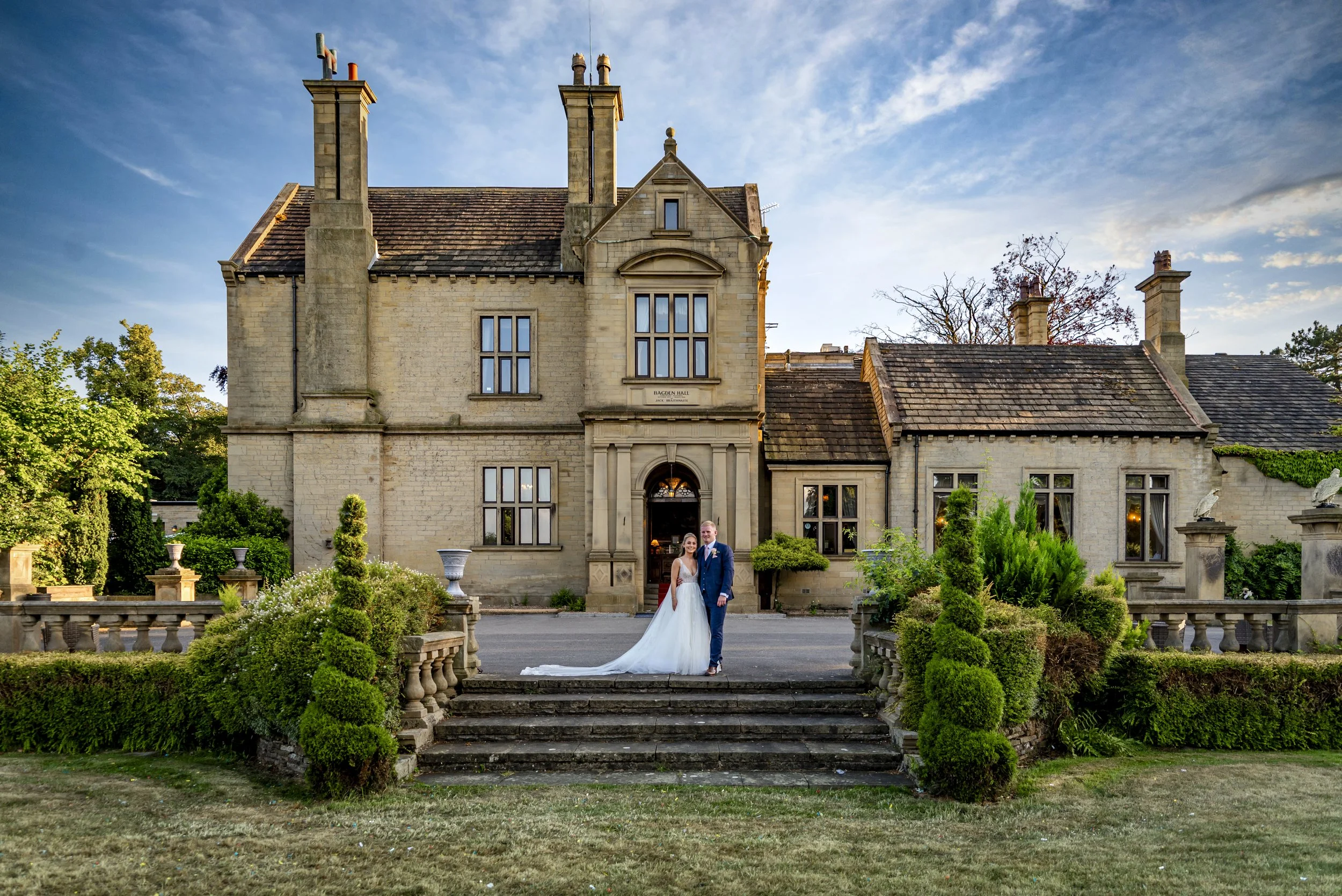 A bride and groom standing on front steps of a historic mansion during sunset, dressed in wedding attire, surrounded by well-maintained garden and greenery.