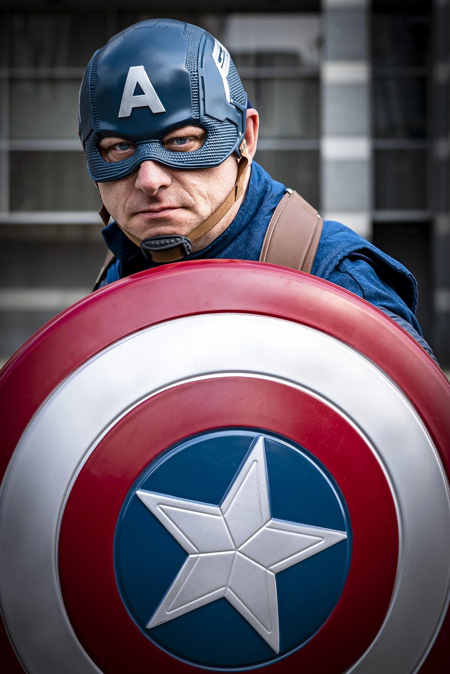 A close-up of a man wearing Captain America's mask and holding his shield. The shield has red, white, and blue concentric circles with a white star in the center. The background appears to be cityscape buildings.