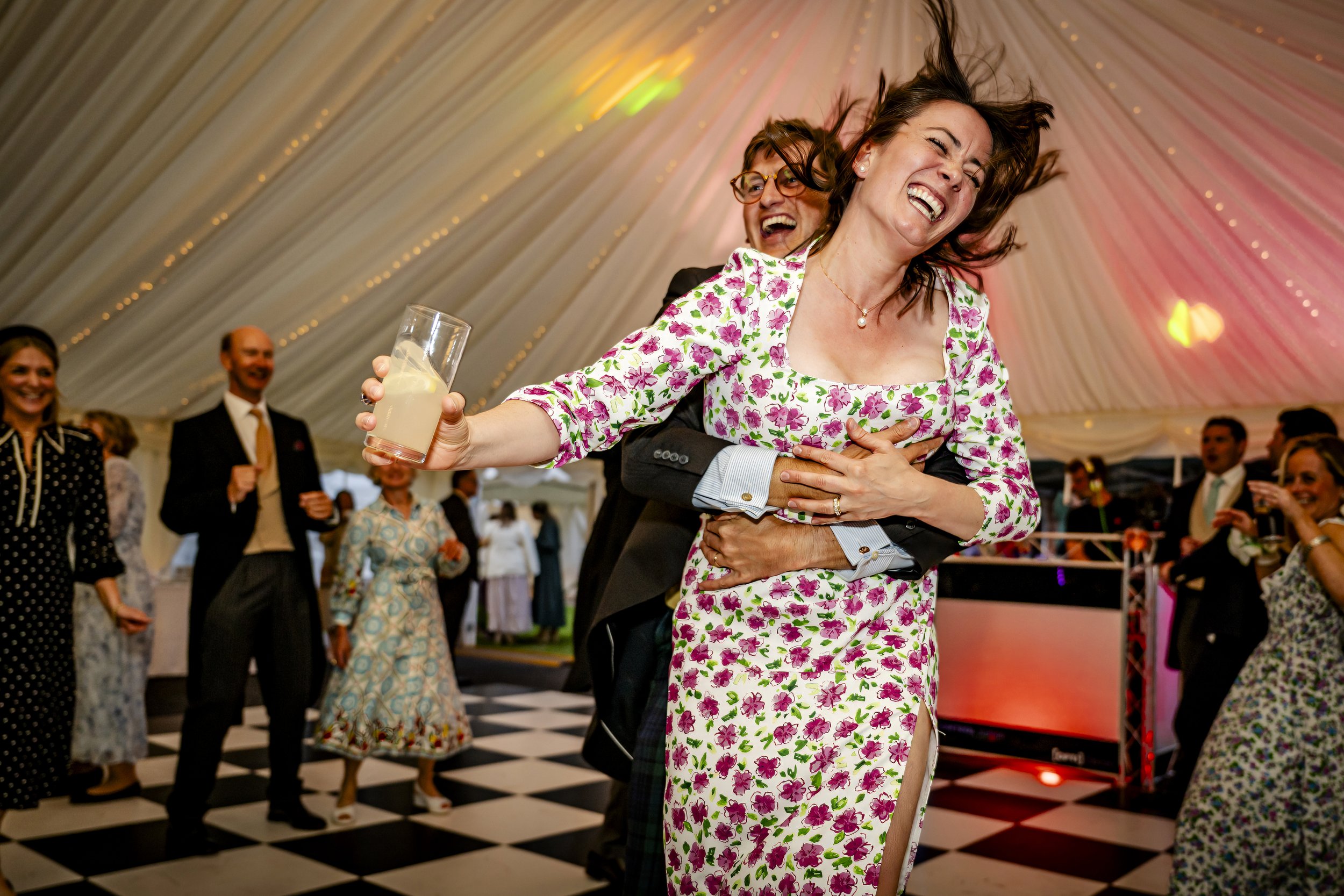 People dancing and celebrating at a wedding reception inside a decorated tent, with a woman in a floral dress and a man in a suit hugging and smiling, holding a drink.