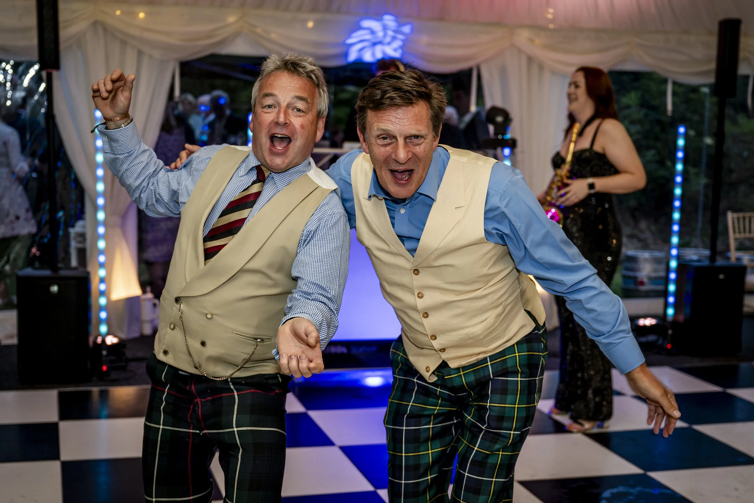 Two men in vintage-style vests and plaid pants dancing and smiling at a lively party with a checkered floor, a stage, and a woman in a black dress playing a saxophone in the background.