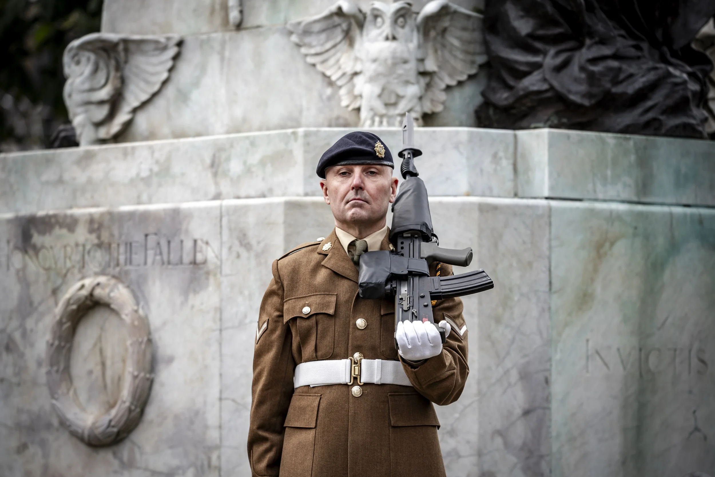 A soldier in uniform standing at attention with a rifle in front of a monument featuring a carved eagle and an inscription.
