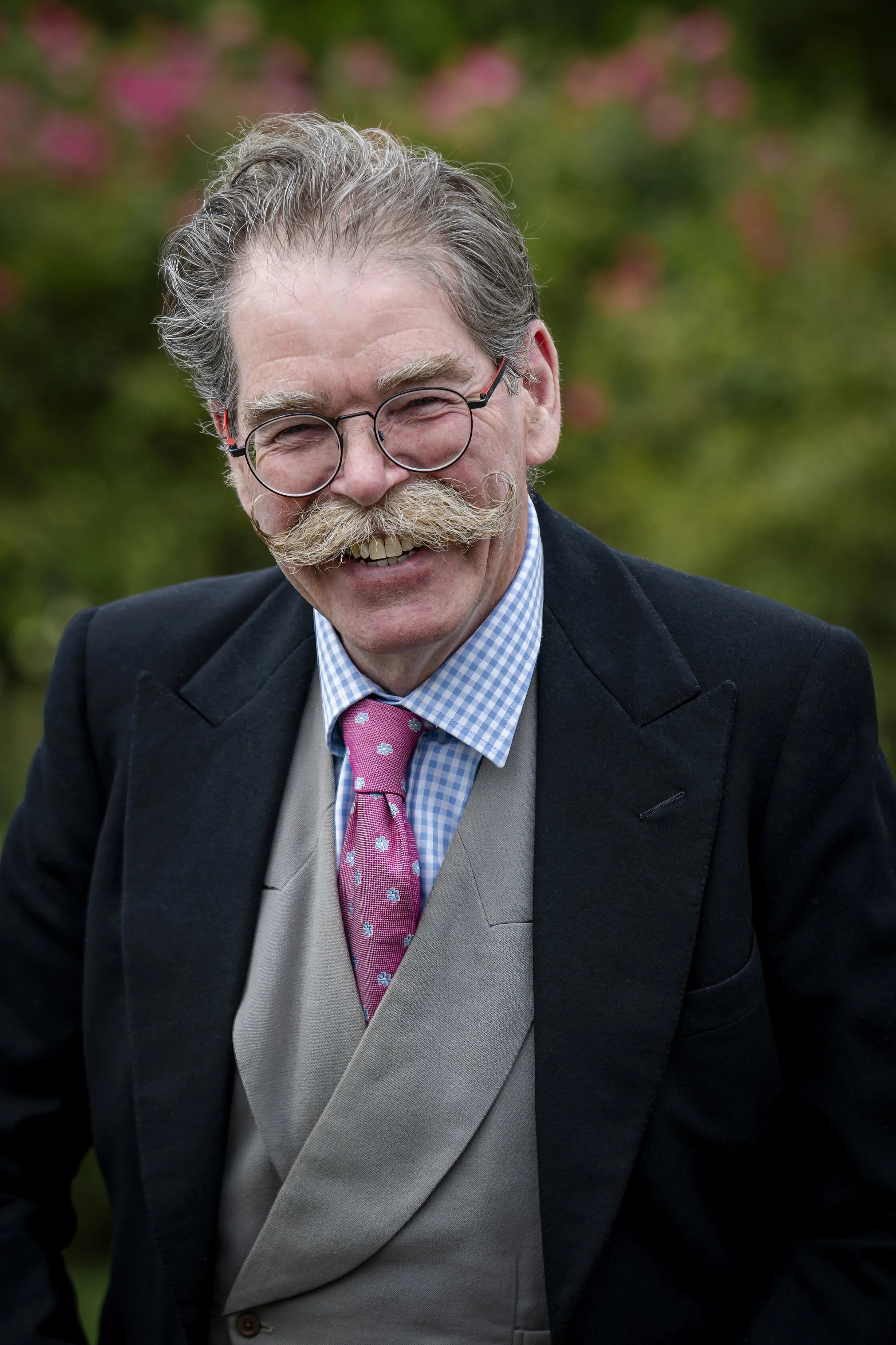 A smiling man with glasses and a mustache, wearing a dark blazer, gray vest, and pink tie, outdoors with blurred green and pink foliage in the background.