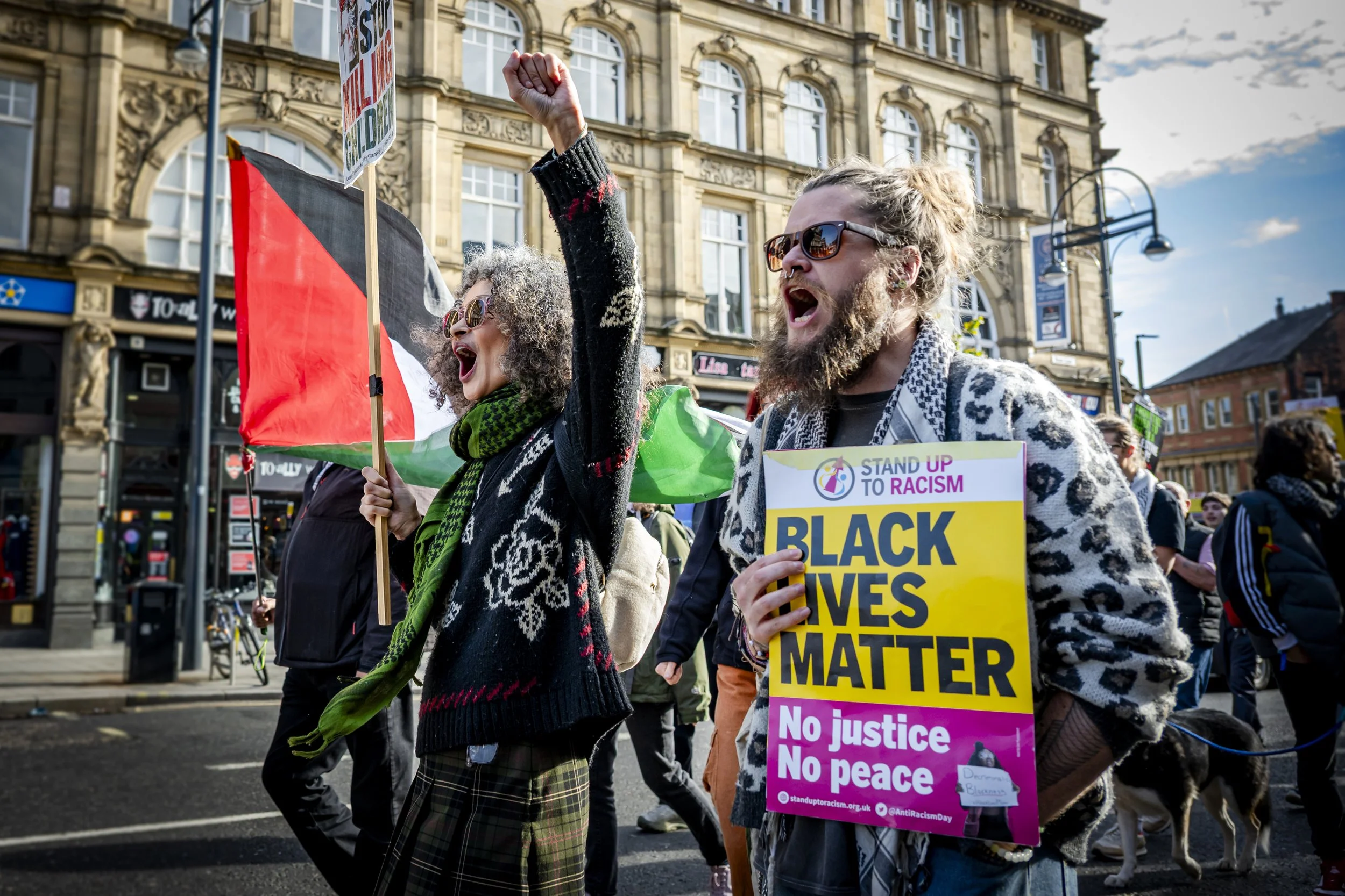 Protesters participating in a Black Lives Matter demonstration on a city street. One person, with a beard and sunglasses, is holding a sign that reads 'BLACK LIVES MATTER' and wearing a patterned shirt. Another person is waving a red, black, and whit