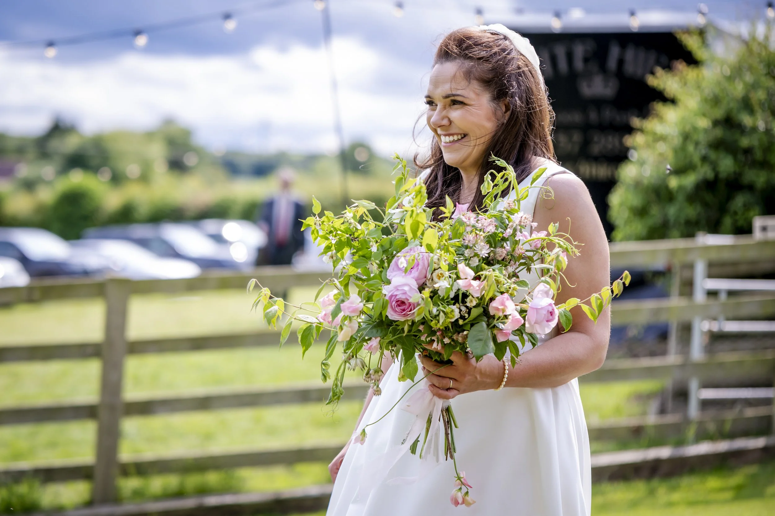 A woman in a white dress holding a large bouquet of pink and white flowers outdoors with a wooden fence behind her and a parking area in the background, smiling happily.