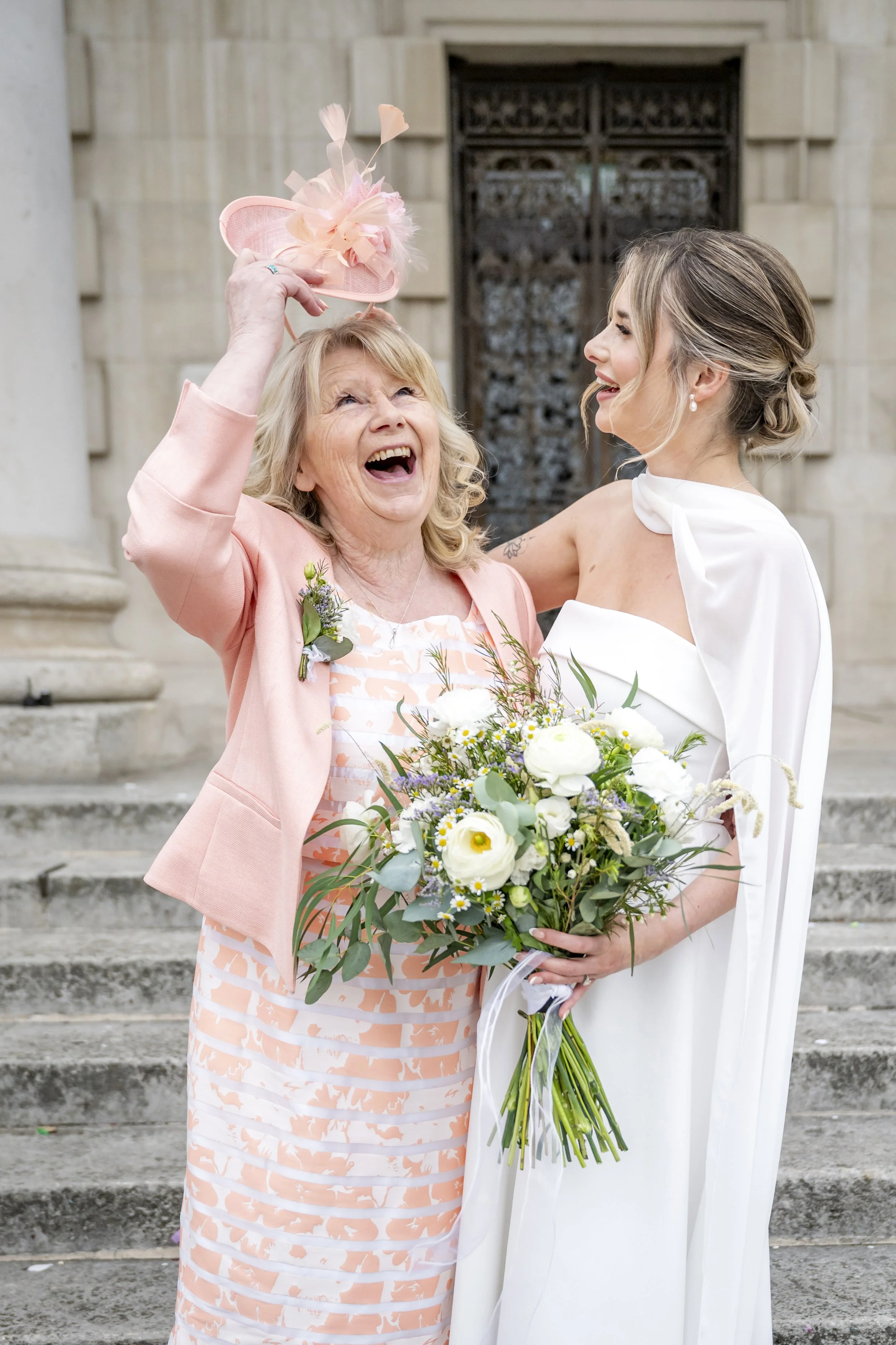 A bride and elderly woman smiling and greeting each other outside a building. The elderly woman is holding a small pink hat with feathers and flowers, and the bride is holding a bouquet of white and purple flowers.