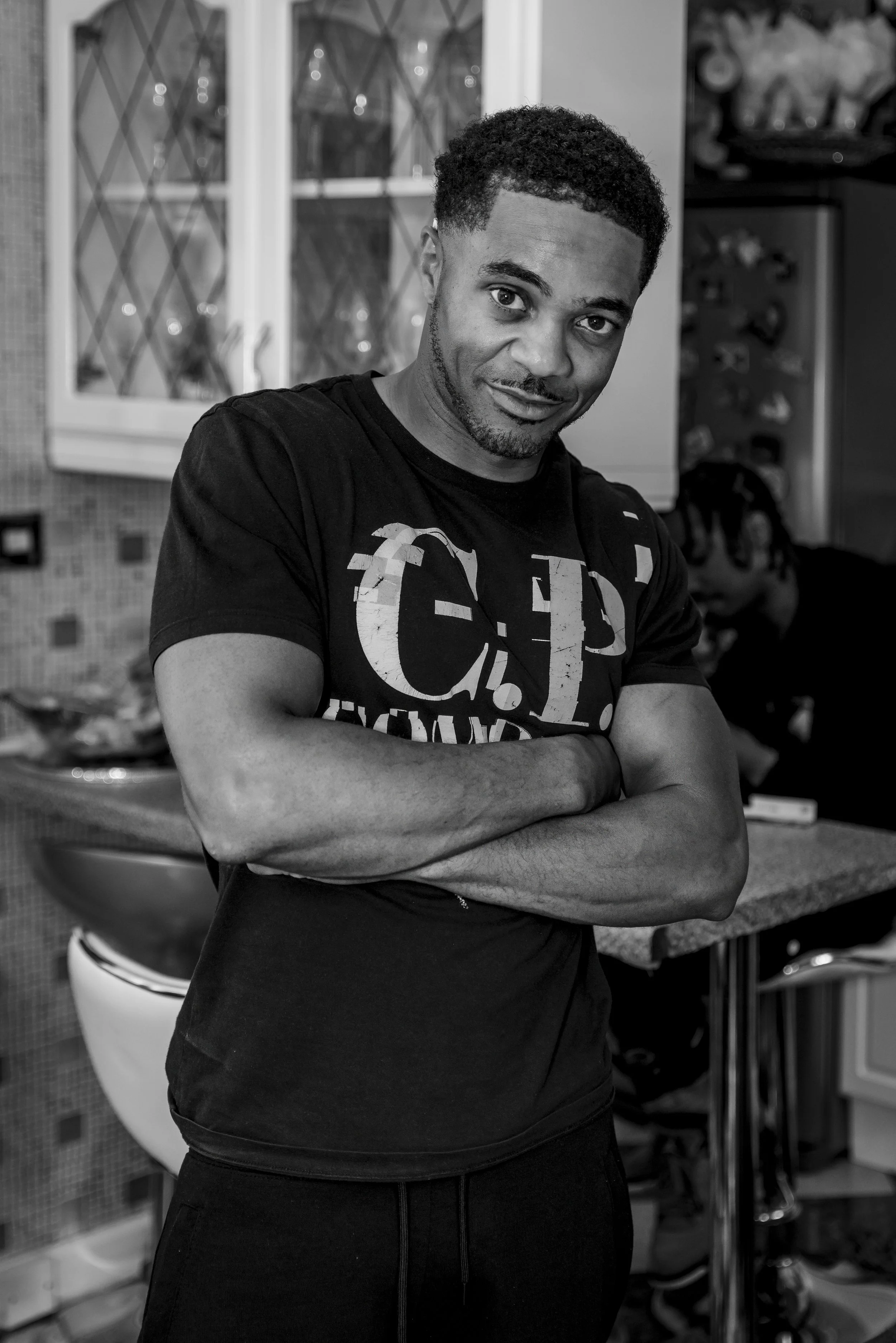 Black and white photo of a man with short curly hair and a beard, standing with arms crossed, smiling slightly, in a kitchen.
