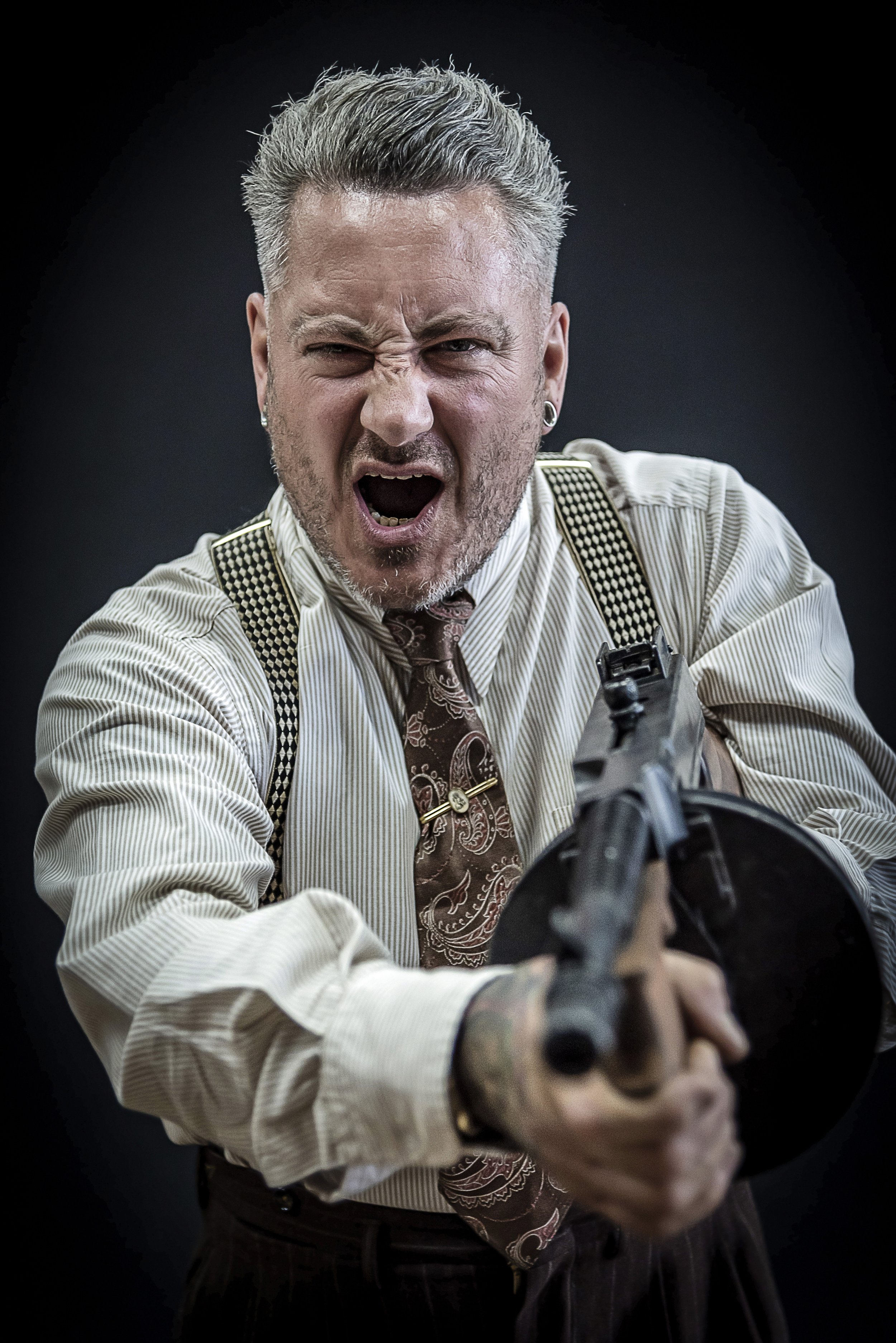 A man with gray hair and a beard, wearing a striped shirt, patterned tie, suspenders, and earrings, is pointing a gun towards the camera with an intense expression on his face, against a dark background.