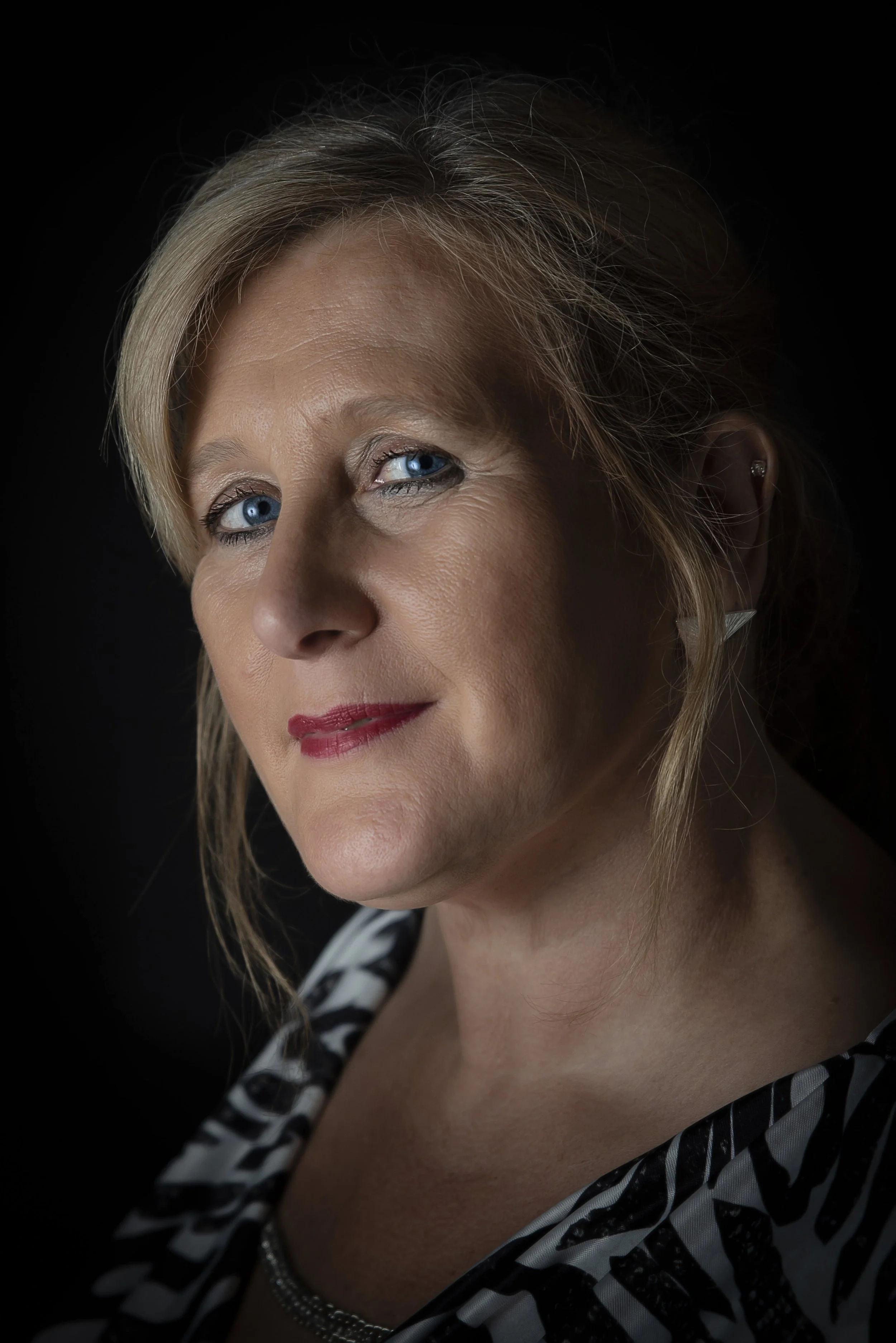 Close-up portrait of a mature woman with blue eyes and blonde hair, wearing red lipstick, earrings, and a black and white patterned top, against a black background.