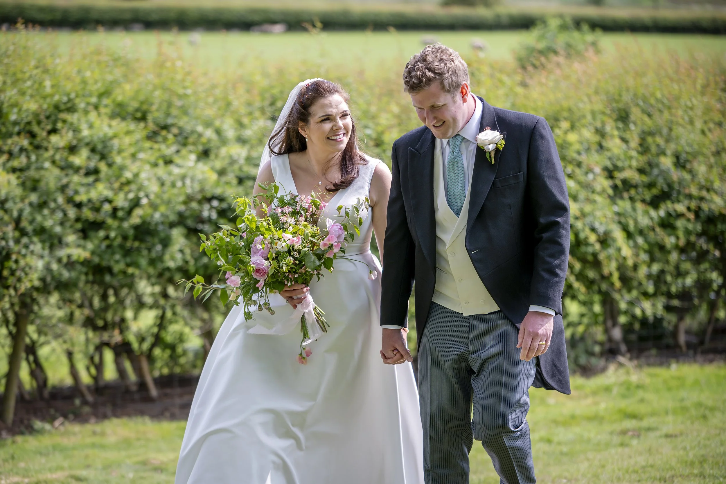 Yorkshire Wedding Photographer - Mike Bettney.
A bride and groom walking on grass outdoors, smiling and holding hands, with the bride holding a bouquet of pink and white flowers, surrounded by green bushes.