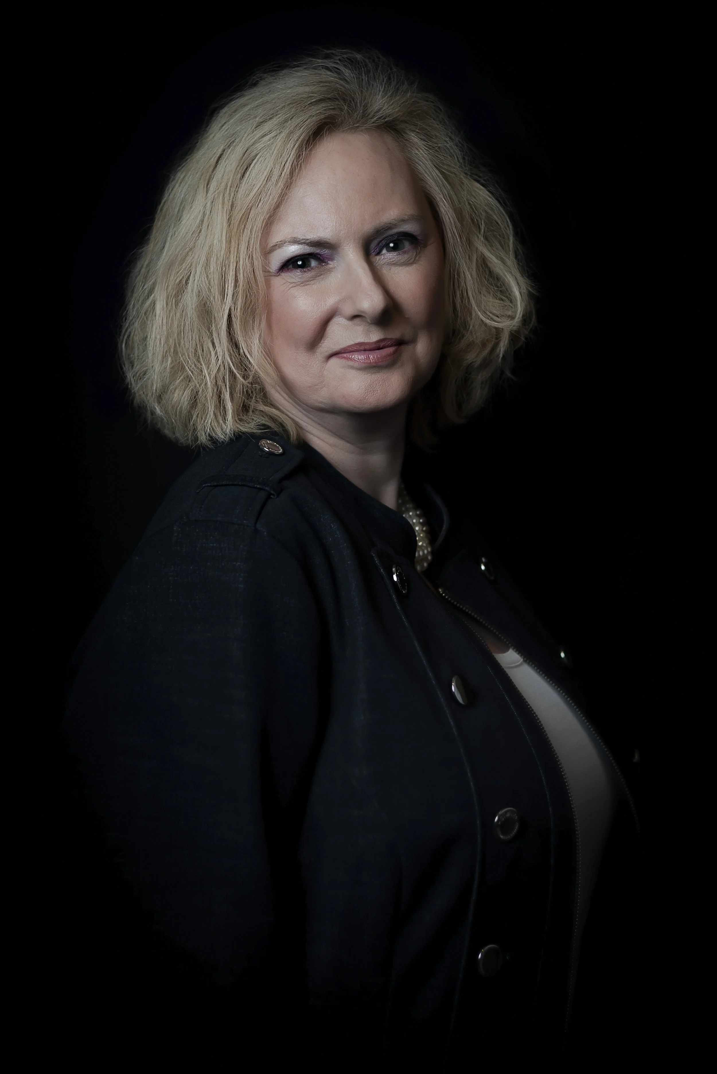 A woman with shoulder-length blonde hair, wearing a black jacket and necklace, posing against a dark background.