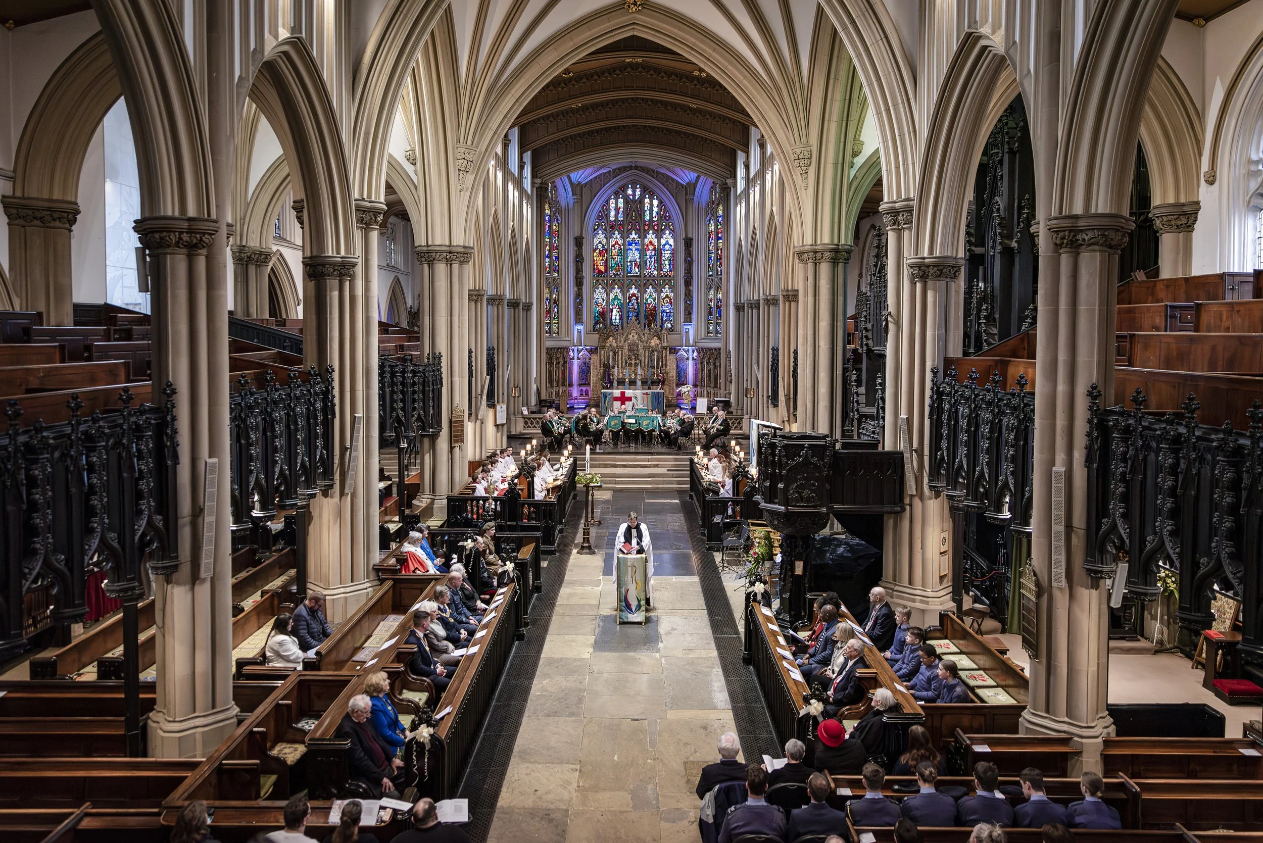 Inside a large Gothic-style cathedral with high vaulted ceilings, stained glass windows, and wooden pews. People are seated and gathered, participating in a religious service or event.