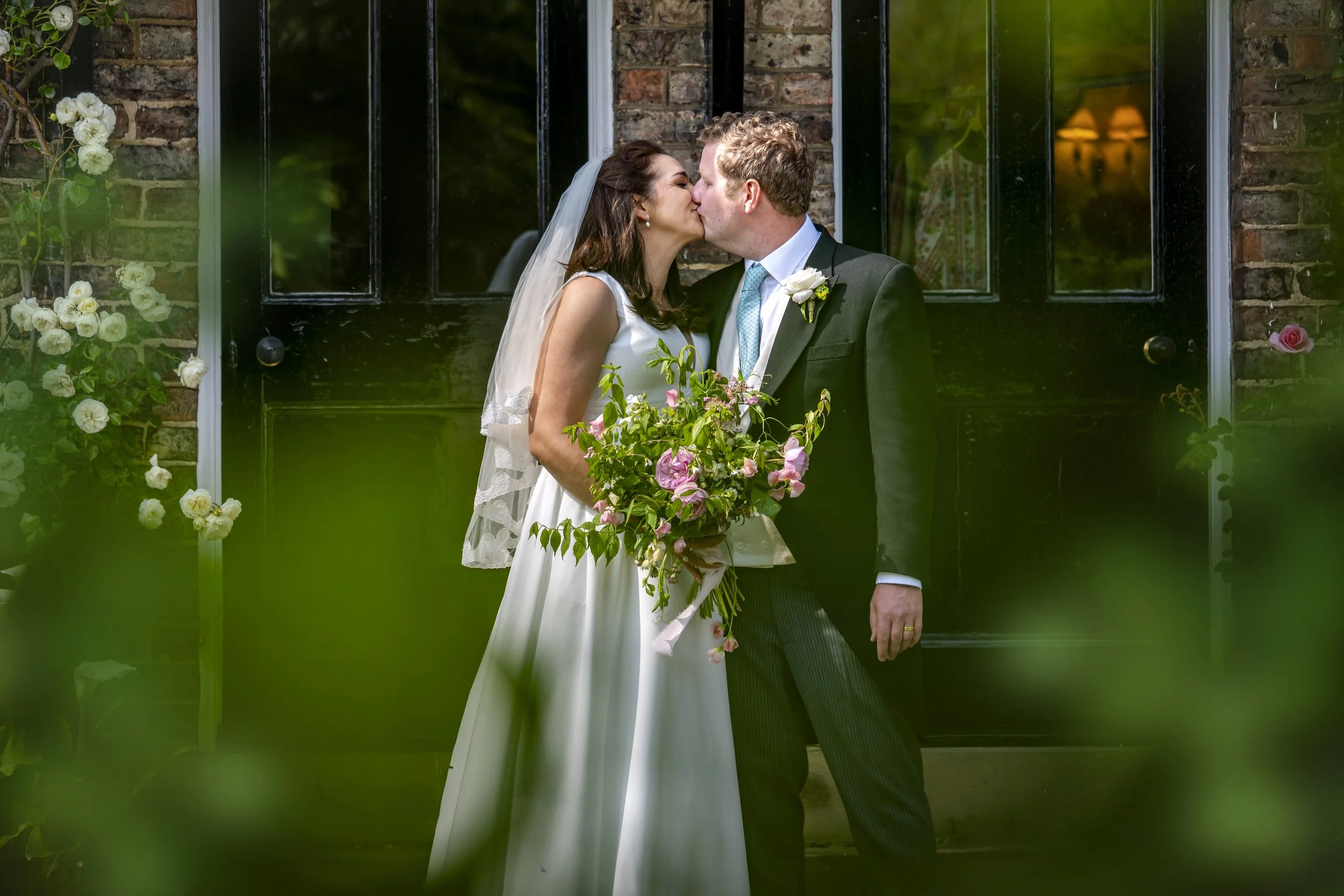 A bride and groom share a kiss outside a brick building on their wedding day. The bride wears a white dress and veil, and holds a bouquet of pink and green flowers. The groom is dressed in a dark suit with a light blue tie and white shirt.
