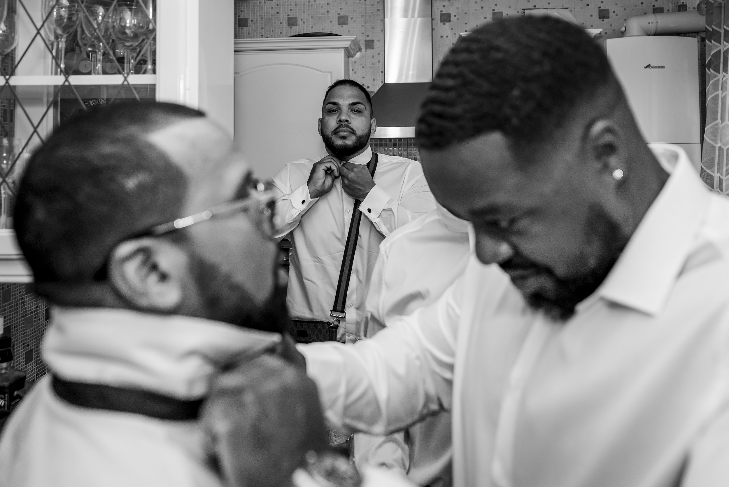 Three men in a kitchen, two of them are focused on a task, one is adjusting a bow tie in the background.