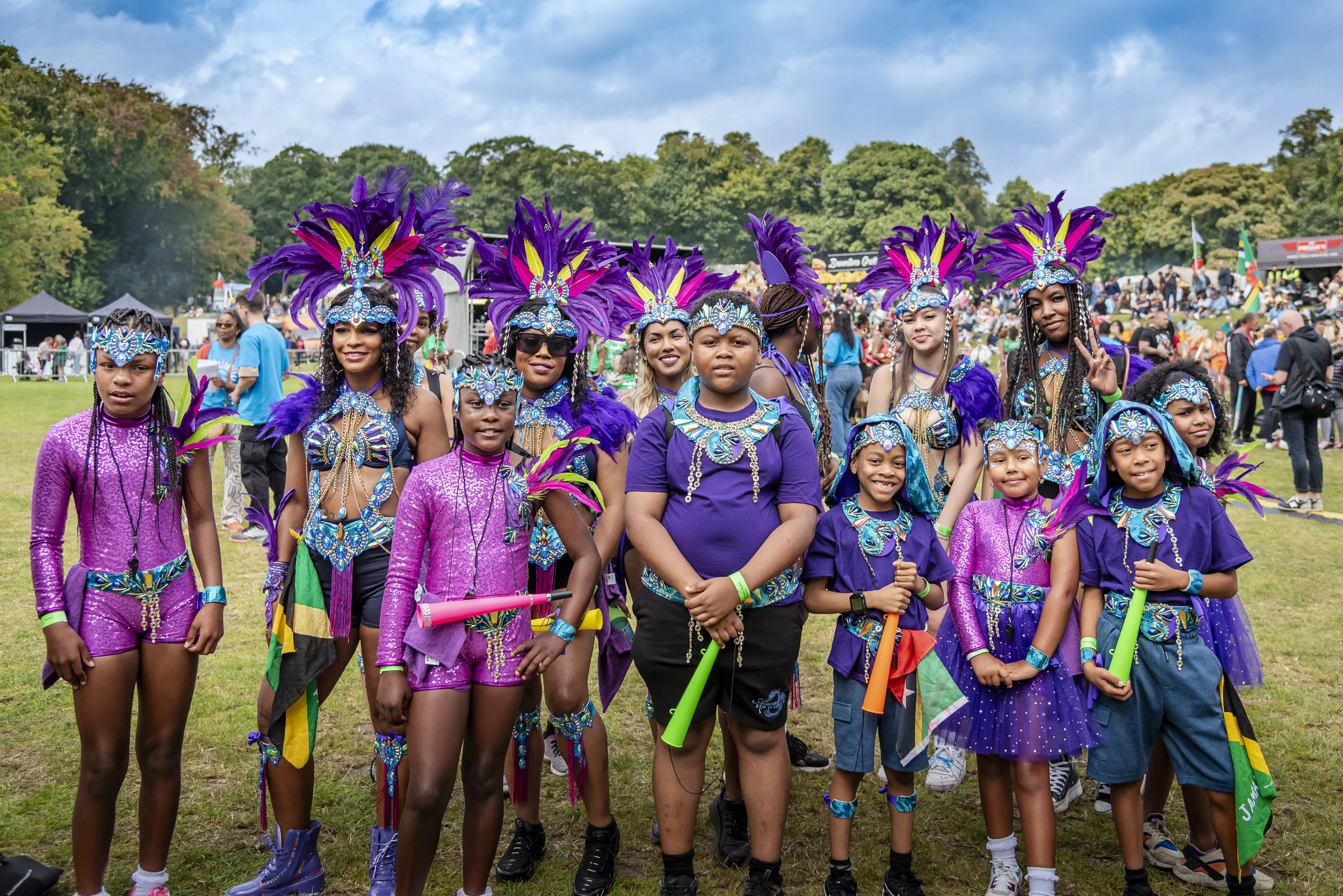 A group of children dressed in vibrant, purple carnival costumes with feathered headdresses, standing outdoors at a festival or celebration, with a crowd and tents in the background.